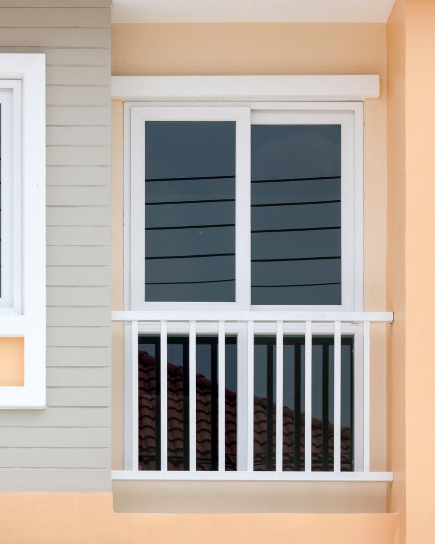 White framed window and balcony on a beige building, with dark tinted glass and vertical railings.