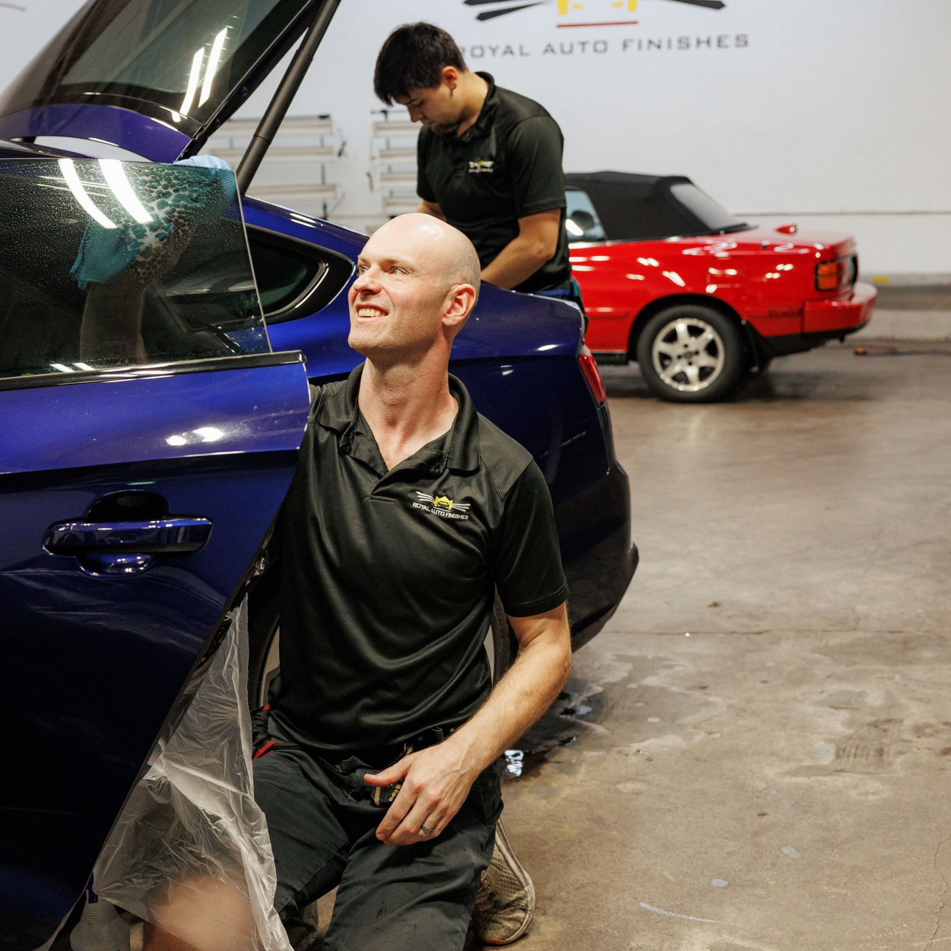 Two people installing tint on a blue car door, in a shop. A red convertible is in the background.