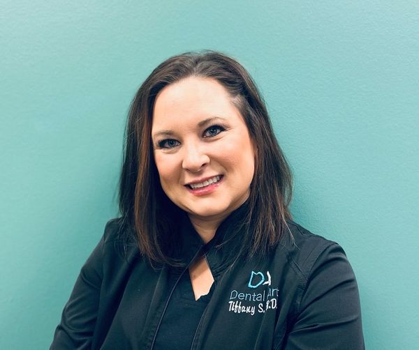 A woman in a black jacket is smiling in front of a blue wall.