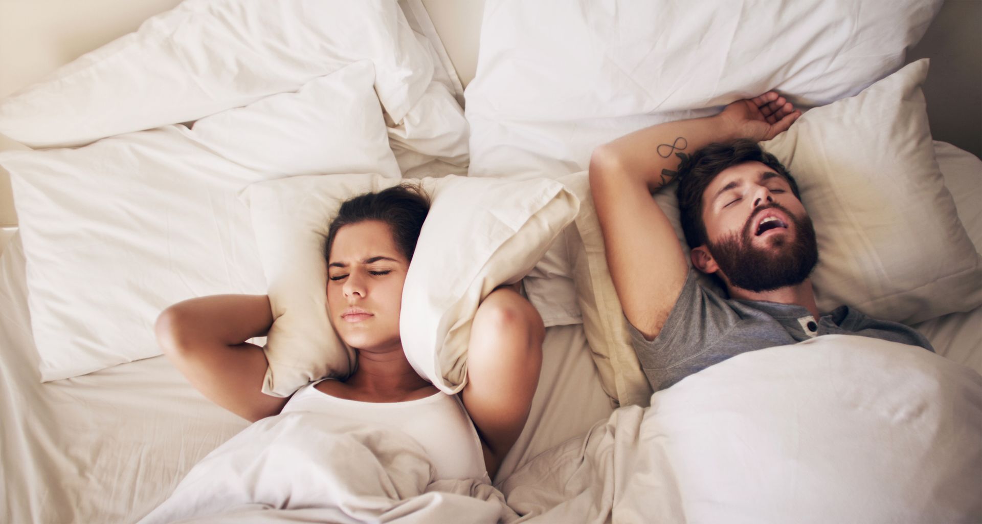 A man is snoring while a woman covers her ears with pillows.