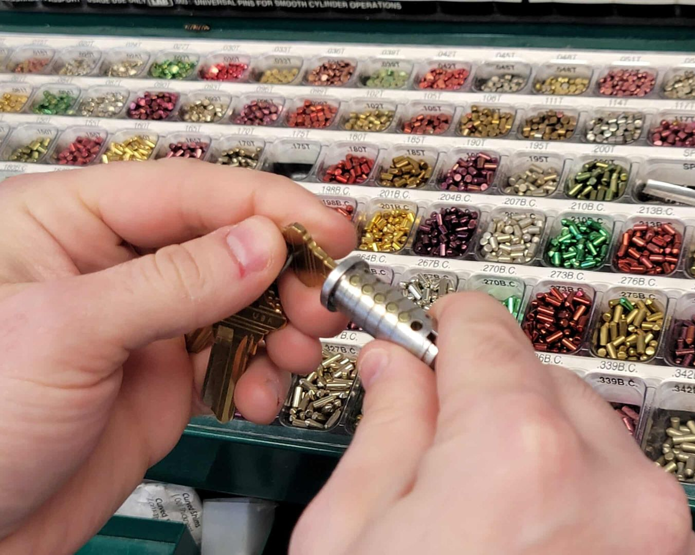 Hands holding a lock cylinder and keys, with a colorful assortment of lock pins in the background.