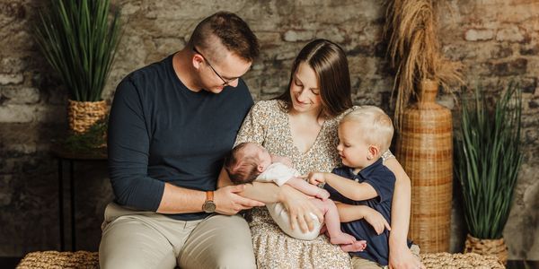 Family with two children poses in front of a brick wall. Mother holds a newborn. Older child reaches for the baby.