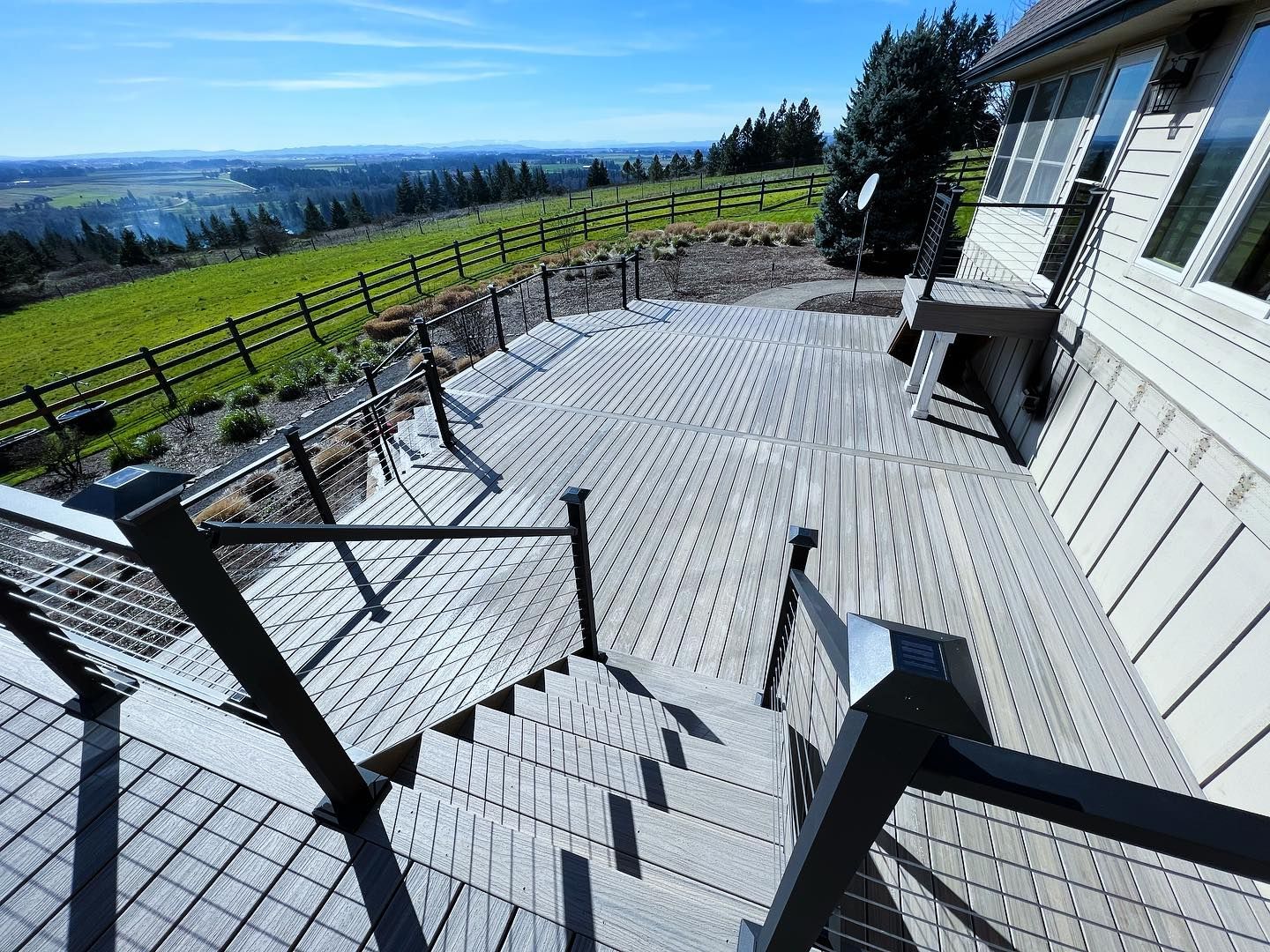 Wooden deck with stairs, cable railings, overlooking a green hillside and blue sky.