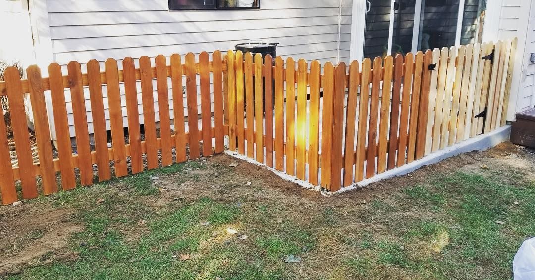 Wooden picket fence with a gate, enclosing a corner of a grassy yard next to a white house.