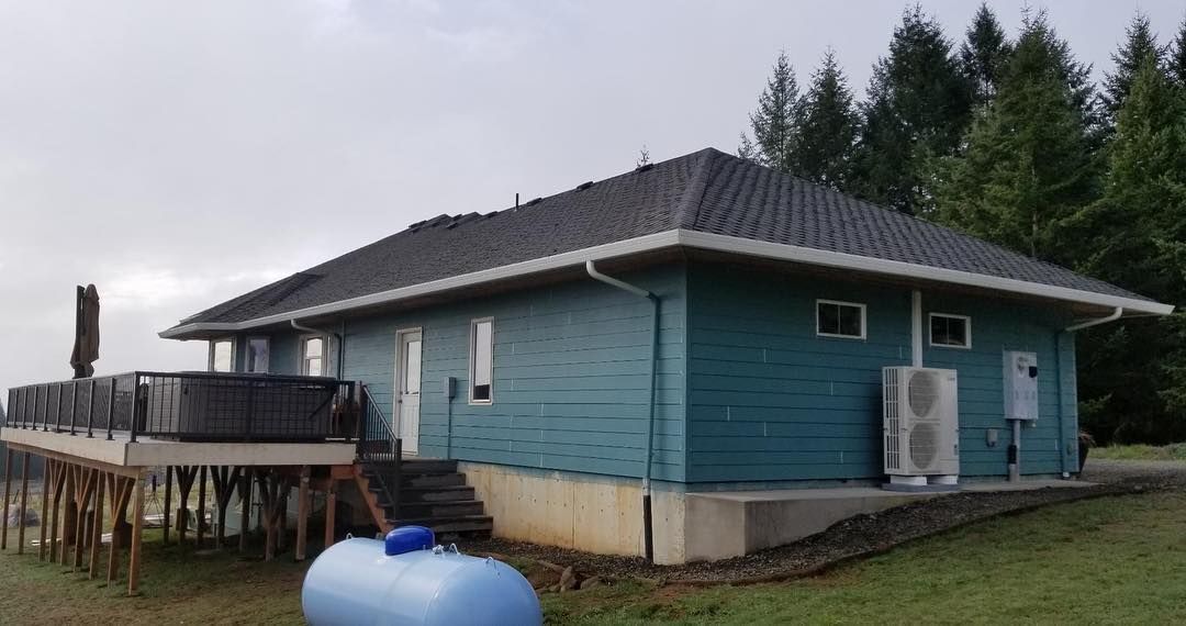Blue house with a black roof and a deck, with a propane tank in the foreground and trees in the background.