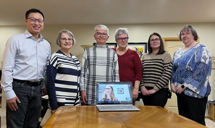 A group of people are standing around a table, and a laptop sits on the table with a Zoom participant.