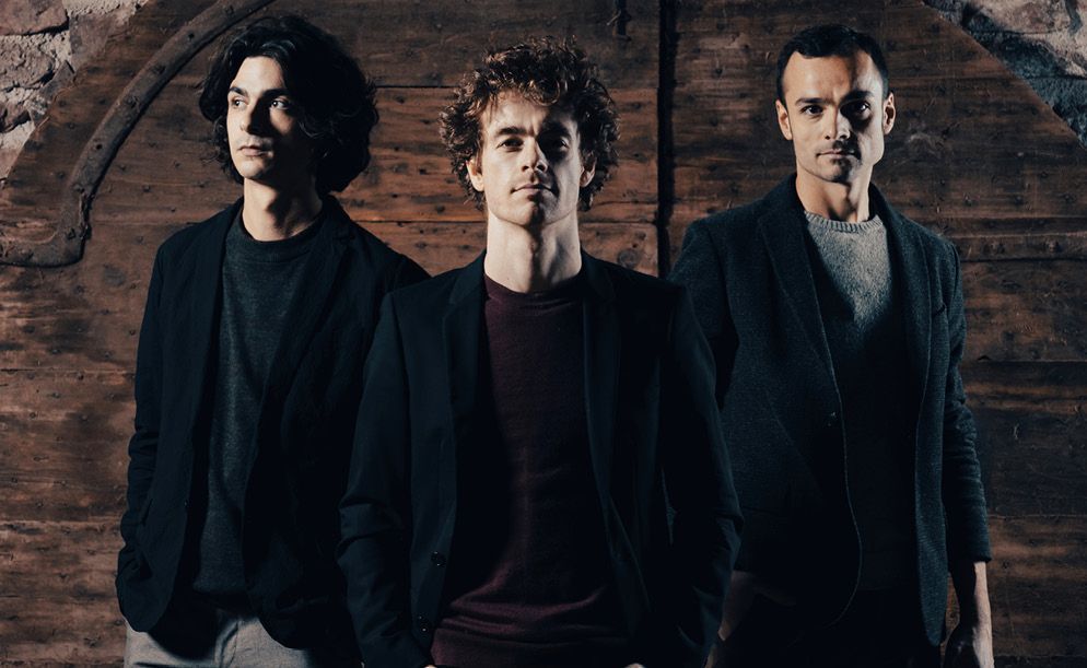 Three men in dark blazers stand in front of a rustic wooden door, posing for a portrait with neutral expressions.