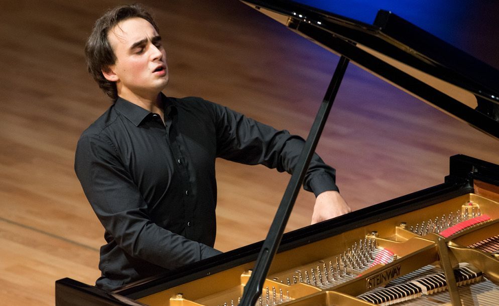 A person wearing a black button-up shirt playing a grand piano with an expressive, concentrated facial expression.
