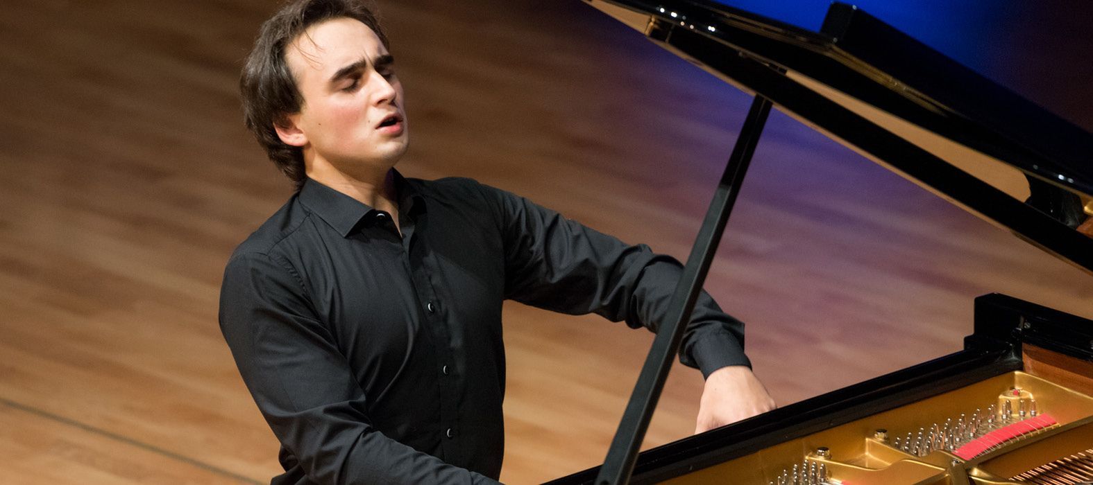 A pianist in a black button-down shirt performs on a grand piano on a wooden stage, eyes closed in concentration.