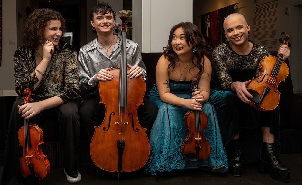 Four musicians sit together indoors, each holding a string instrument; they are dressed in formal, textured attire.