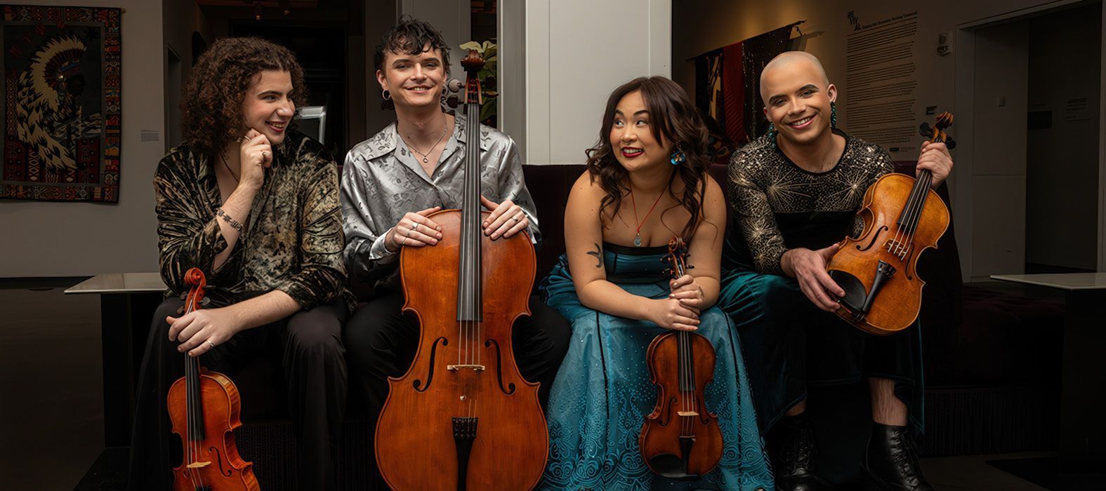 Four musicians sit together in a room, holding their instruments: a cello, two violins, and a viola.