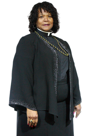 Woman in black clerical attire, smiling slightly. Gold chain necklace. Standing.