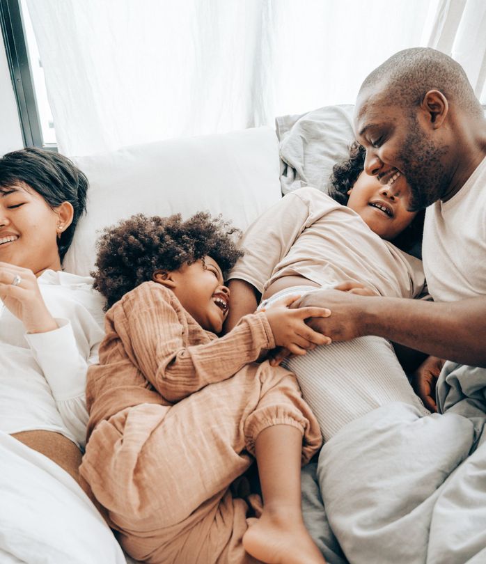 Family relaxing together on a bed, smiling and cuddling under white bedding.