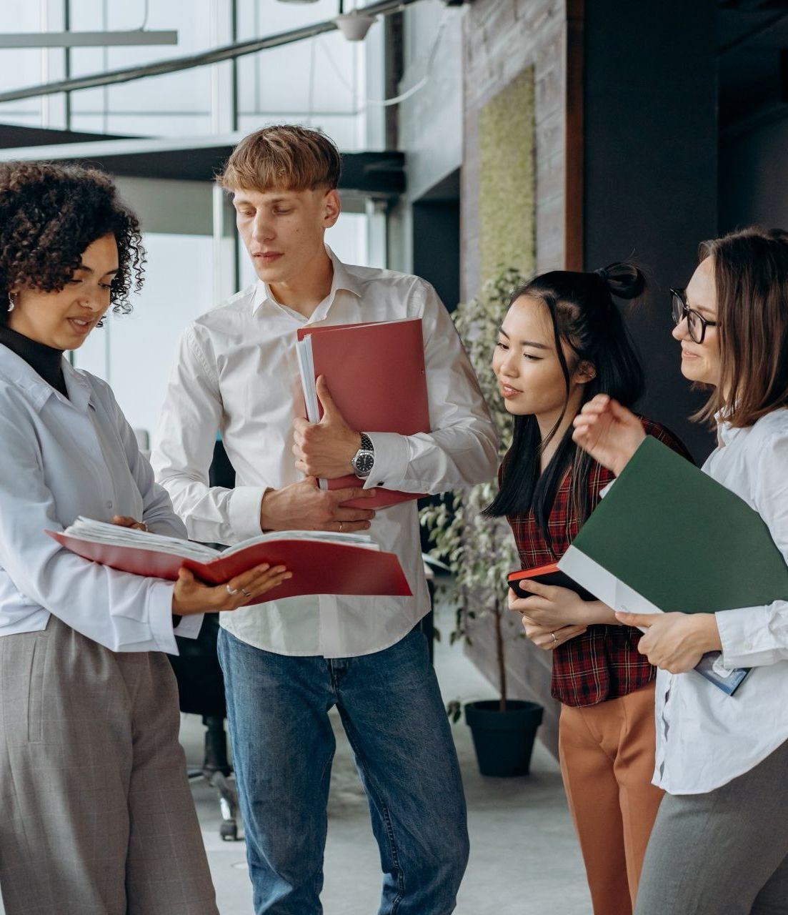 Four coworkers in a modern office discussing documents and holding folders.