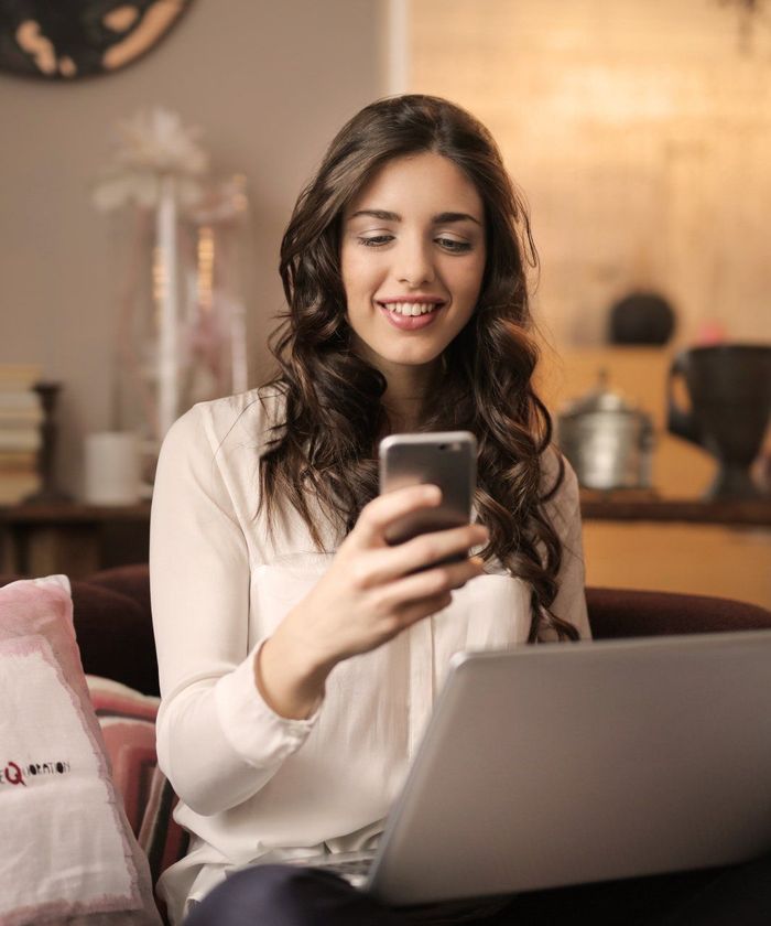 Woman smiling at a laptop while holding a smartphone in a cozy indoor setting