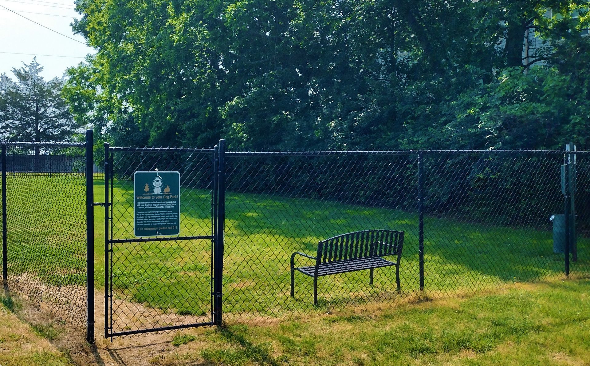 A black metal fenced-in dog park with a wooden bench on the grass, a small sign on the gate, and trees in the background.