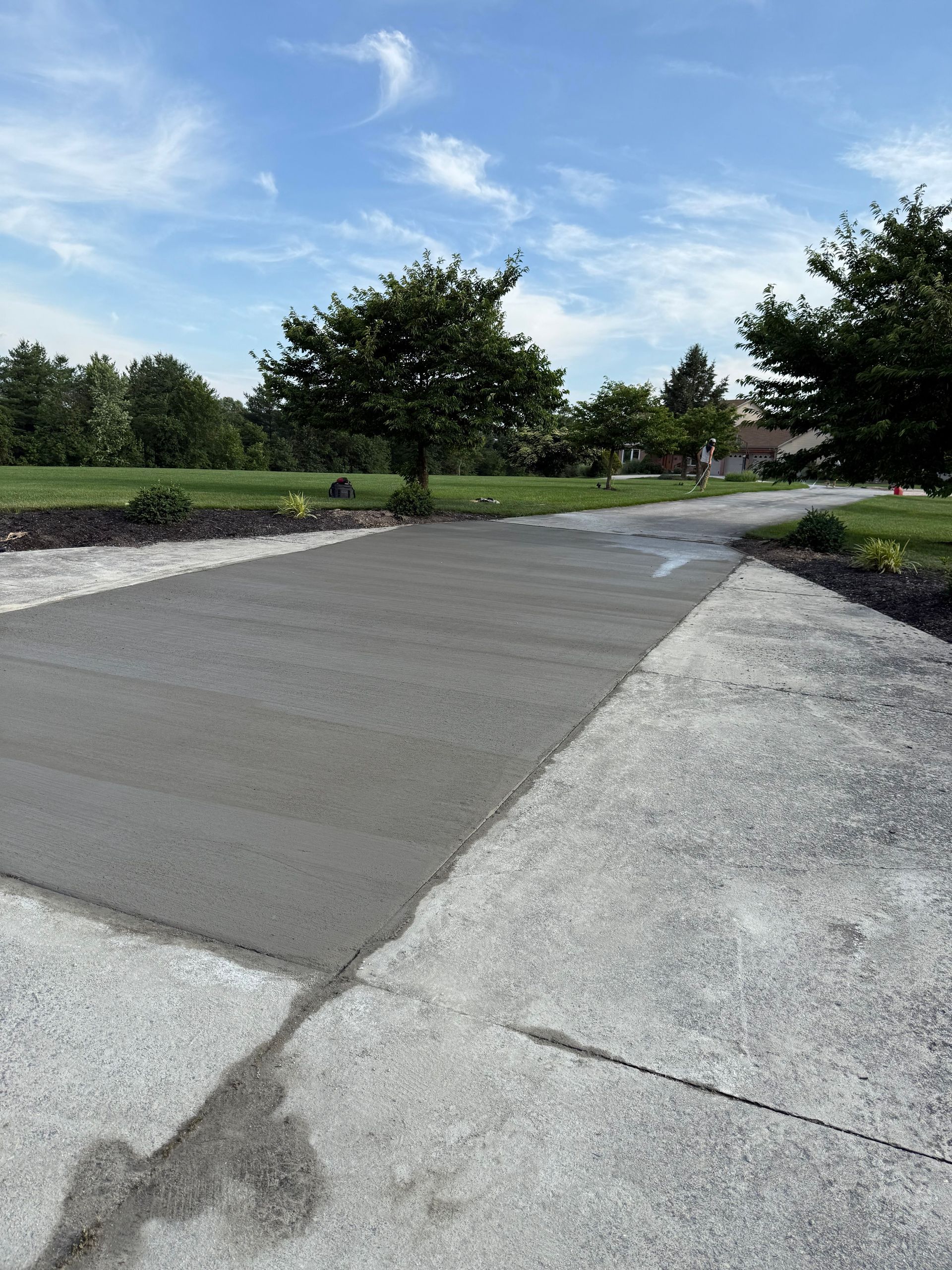 Fresh concrete driveway section with existing driveway, trees, and blue sky.