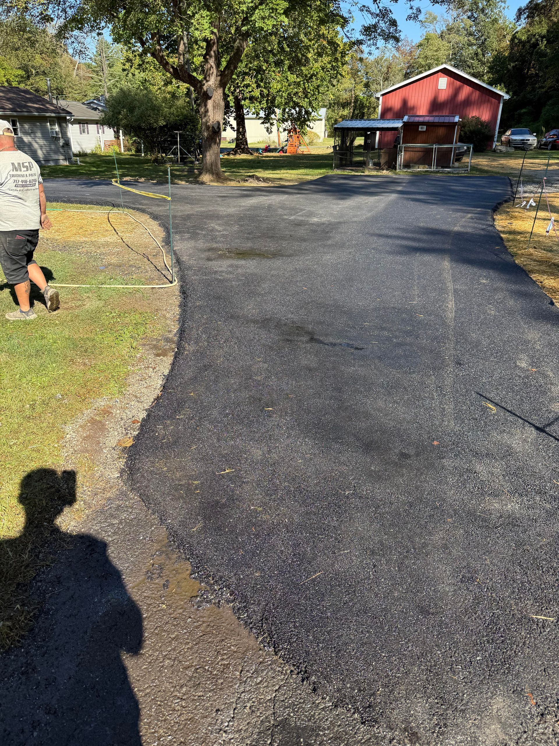 Asphalt driveway with a person standing on grass, red building in the background. Sunny day.