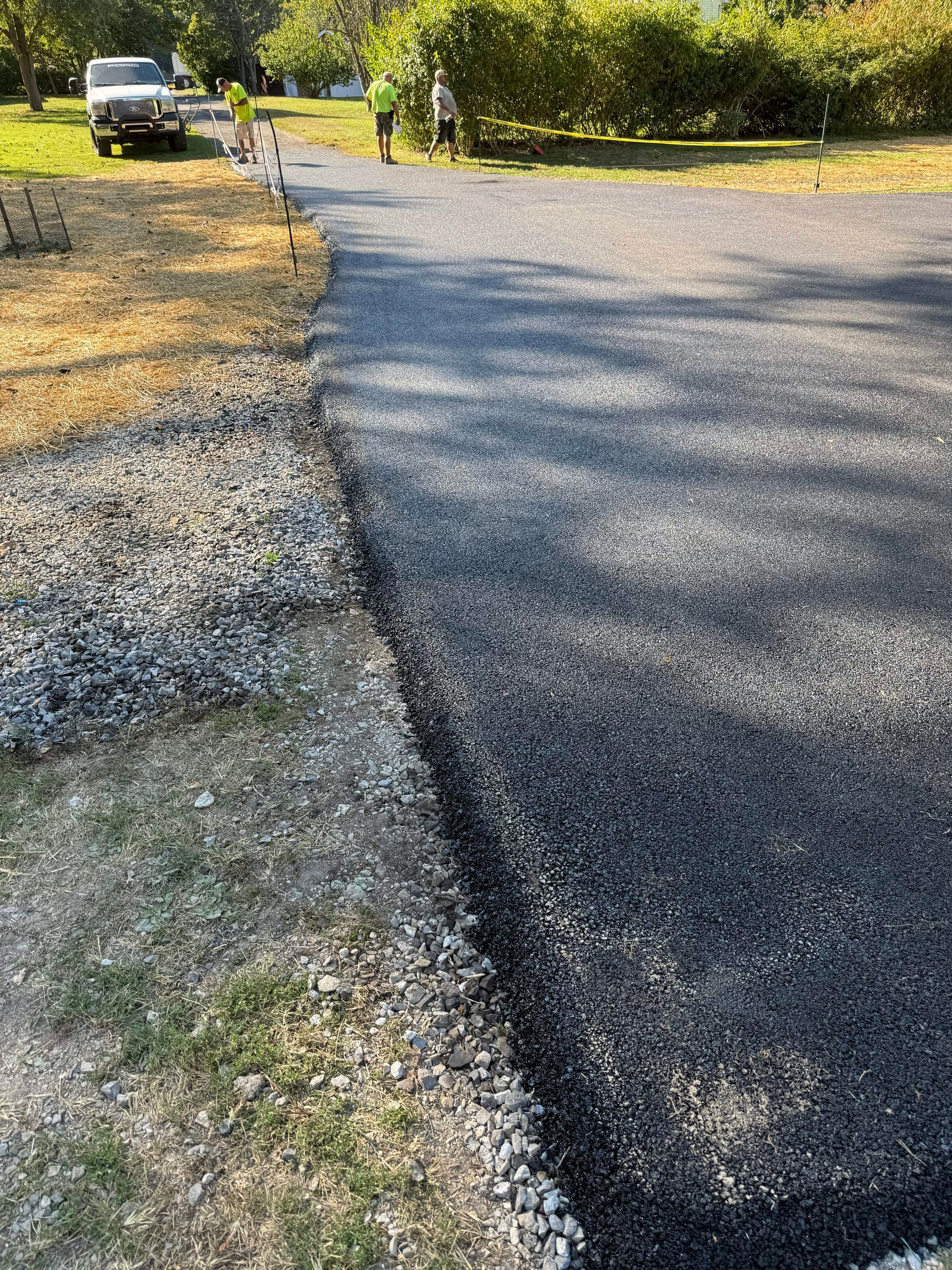 Asphalt driveway being installed; workers in safety vests; gravel edge next to grass.