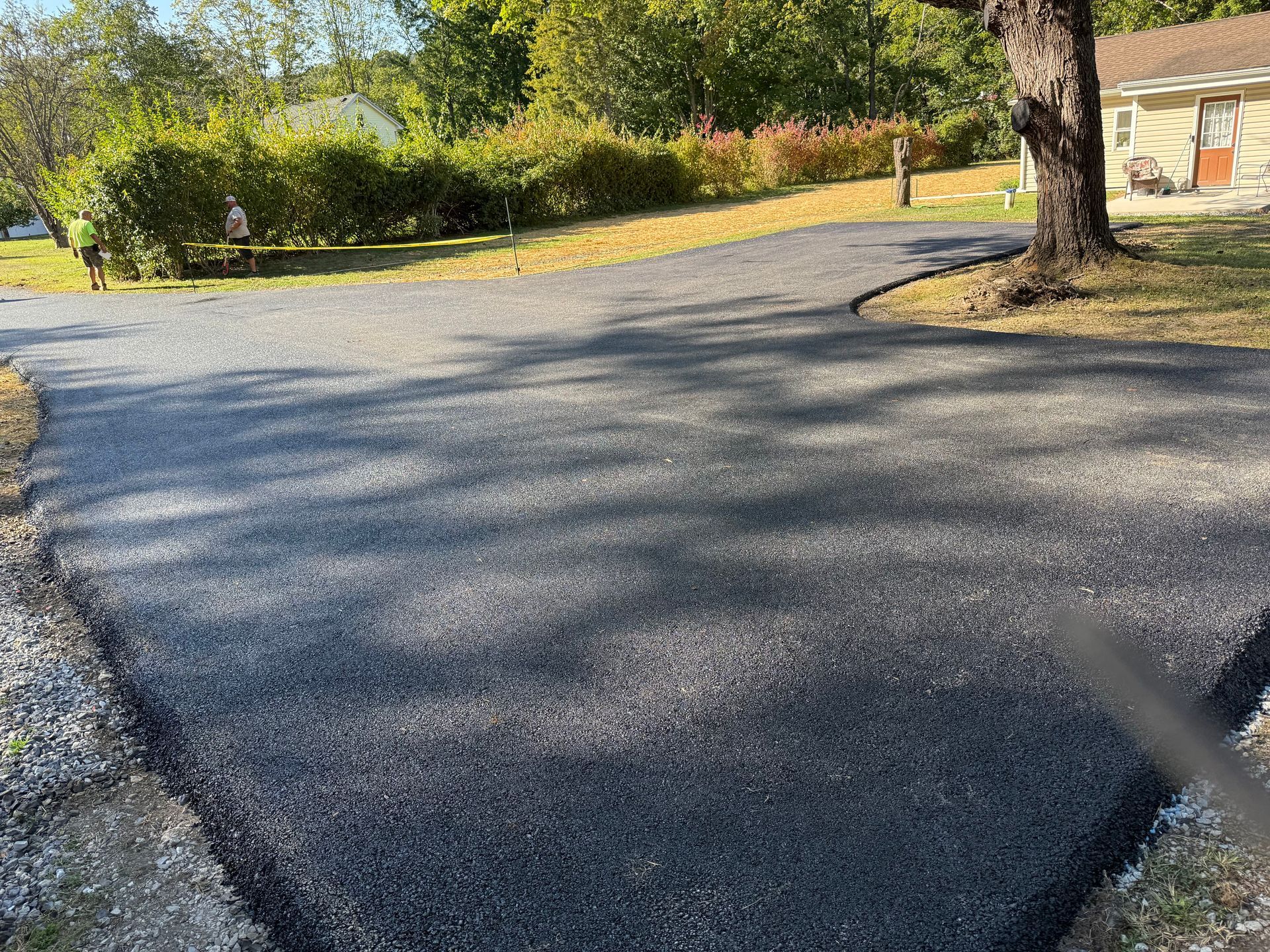 Newly paved asphalt driveway curves towards a house, trees, and foliage in daylight.
