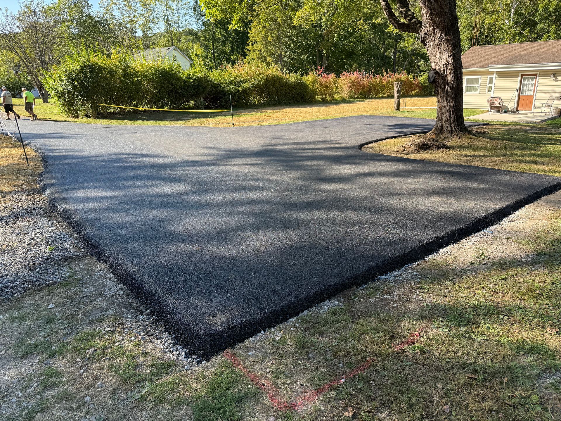 New asphalt driveway with gravel border. Green grass, trees, and a house in the background.