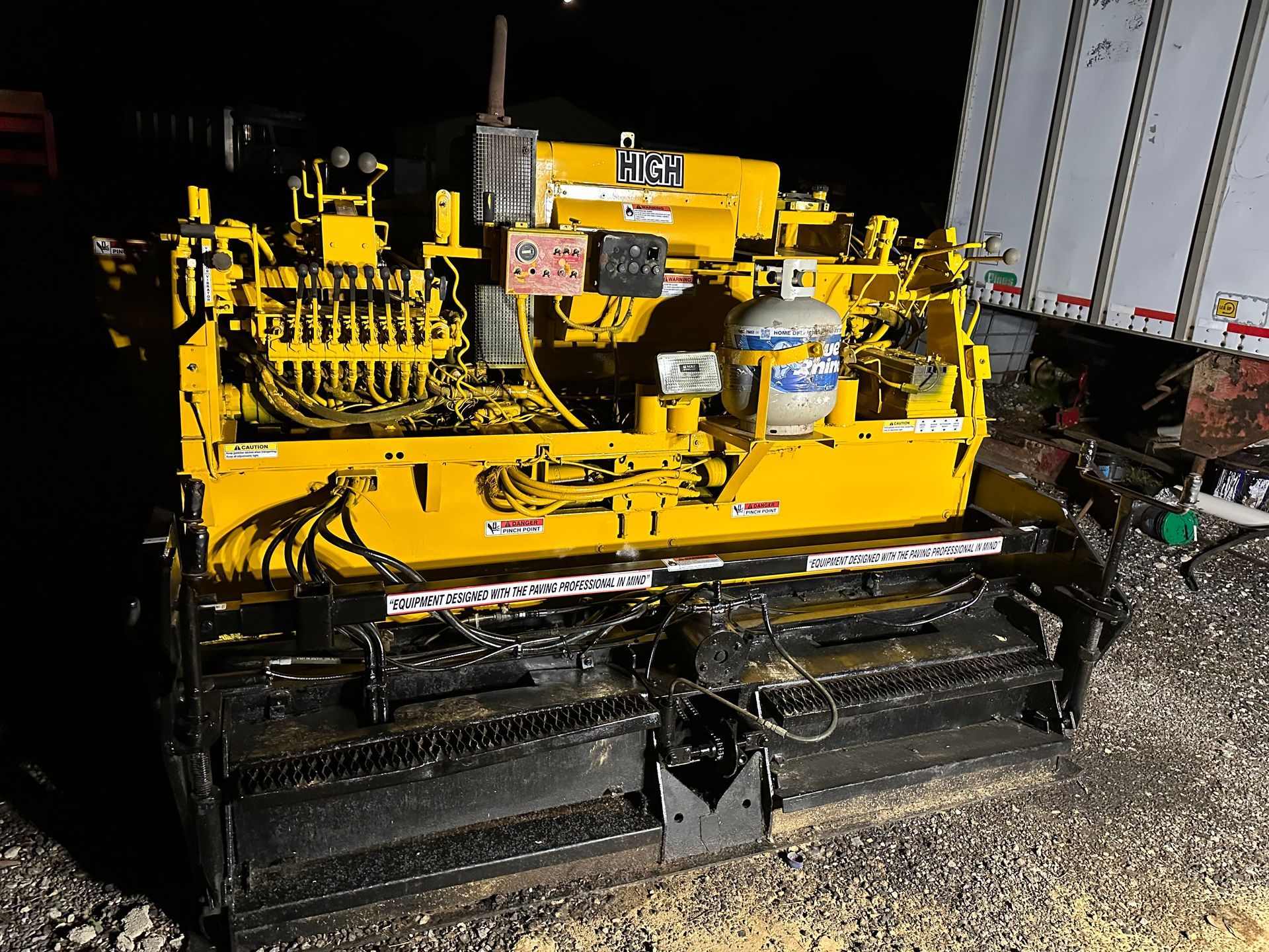 Yellow asphalt paving machine in a dark setting, with a propane tank attached.