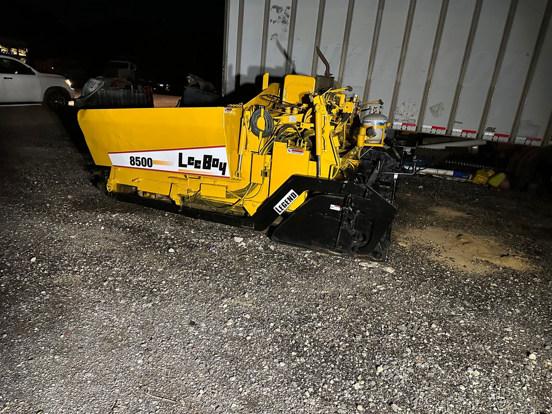 Yellow road maintenance machine damaged, resting on gravel near a semi-trailer.