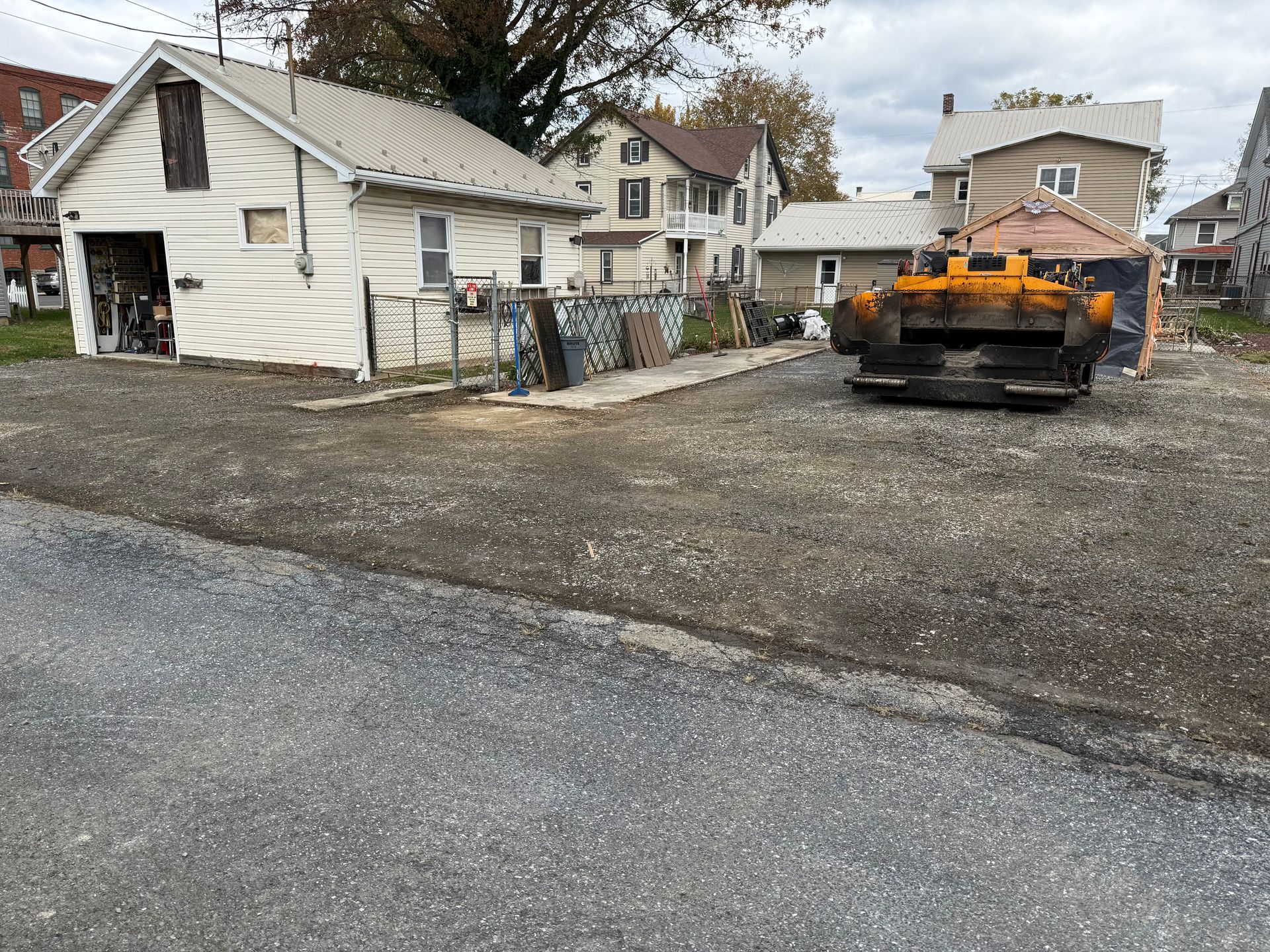 Asphalt paving equipment and work site with buildings in the background. Gravel and asphalt surfaces.