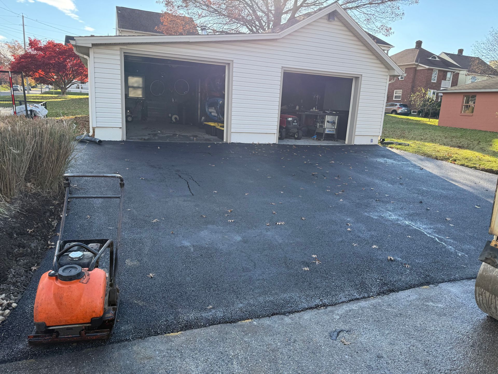 Freshly paved black asphalt driveway in front of a white two-car garage. An orange compactor sits on the asphalt.