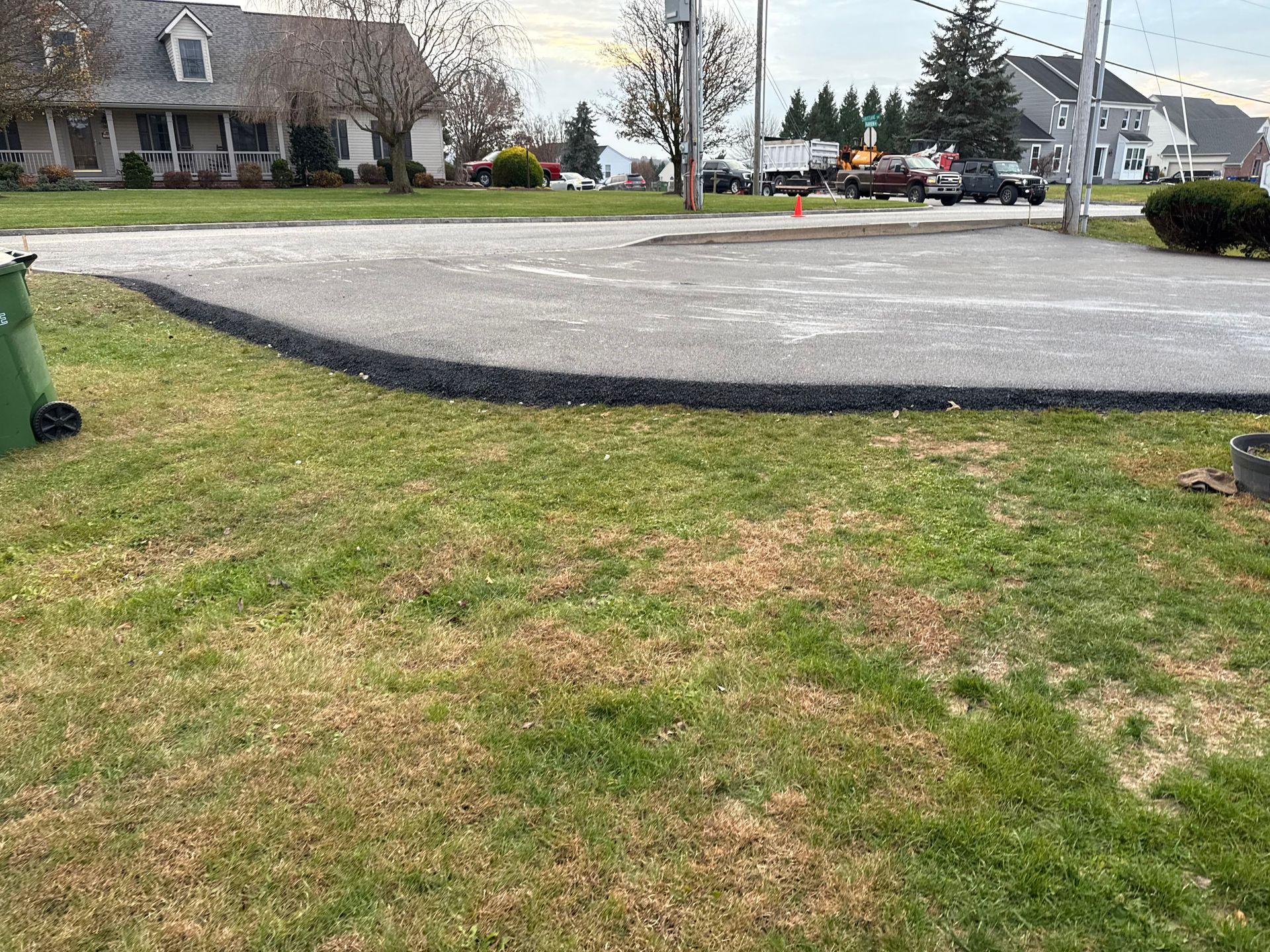 Grassy lawn with a dark asphalt curve, leading to a street with houses and a utility pole.