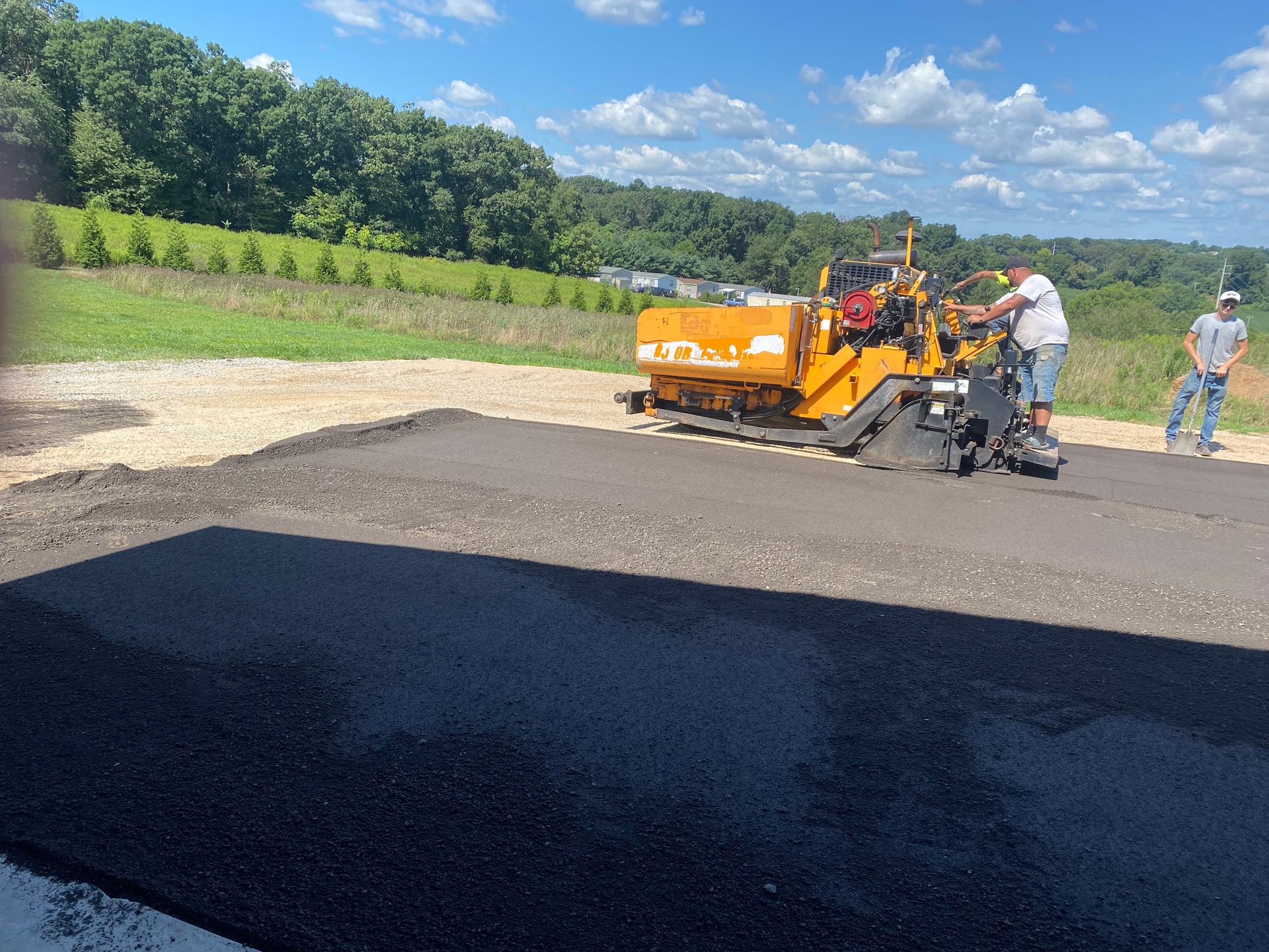 Asphalt paving machine laying blacktop; two workers near machine, sunny day.
