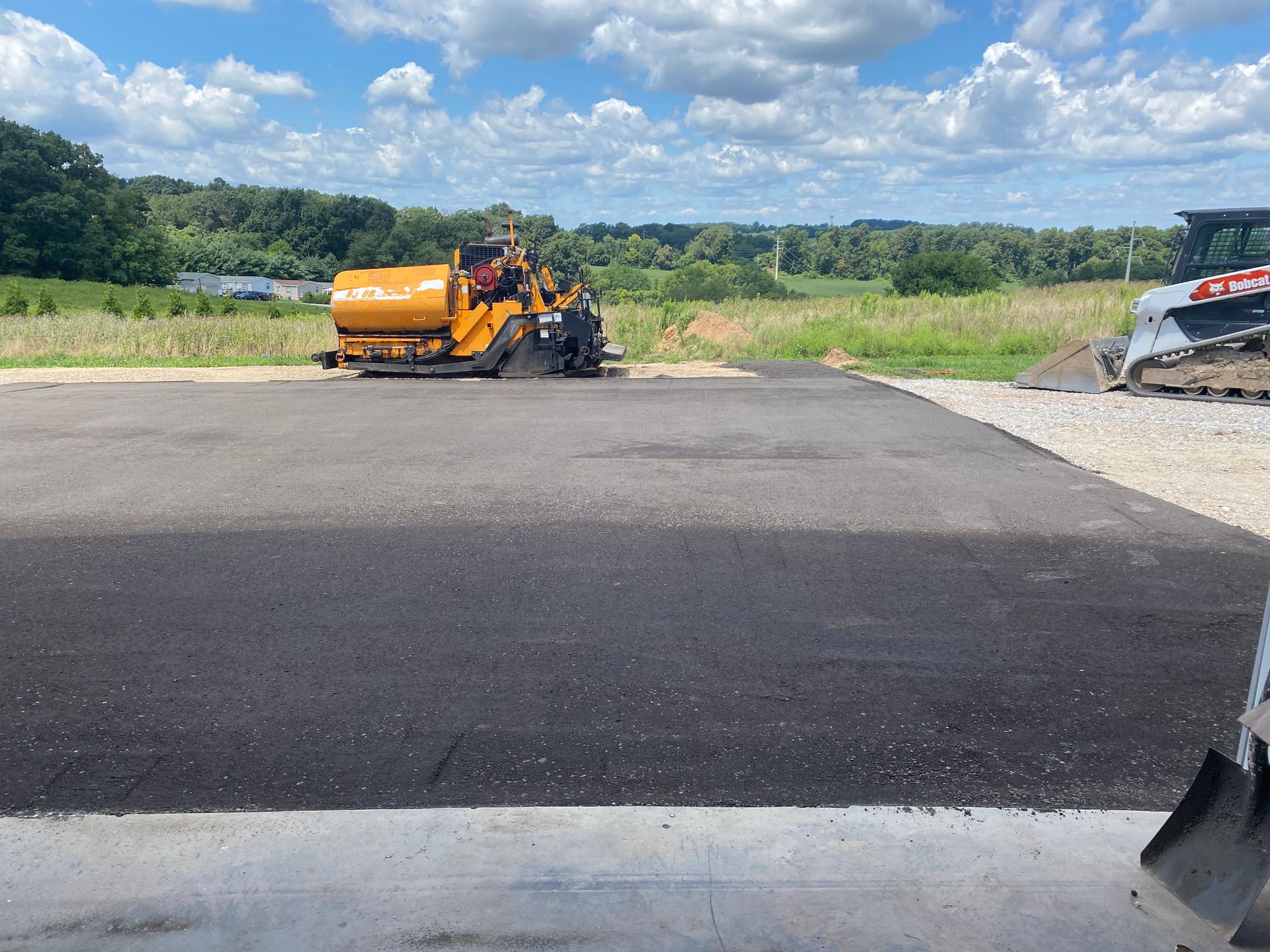 Asphalt paving in progress on a sunny day. Paver machine laying asphalt on new surface; Bobcat in the background.
