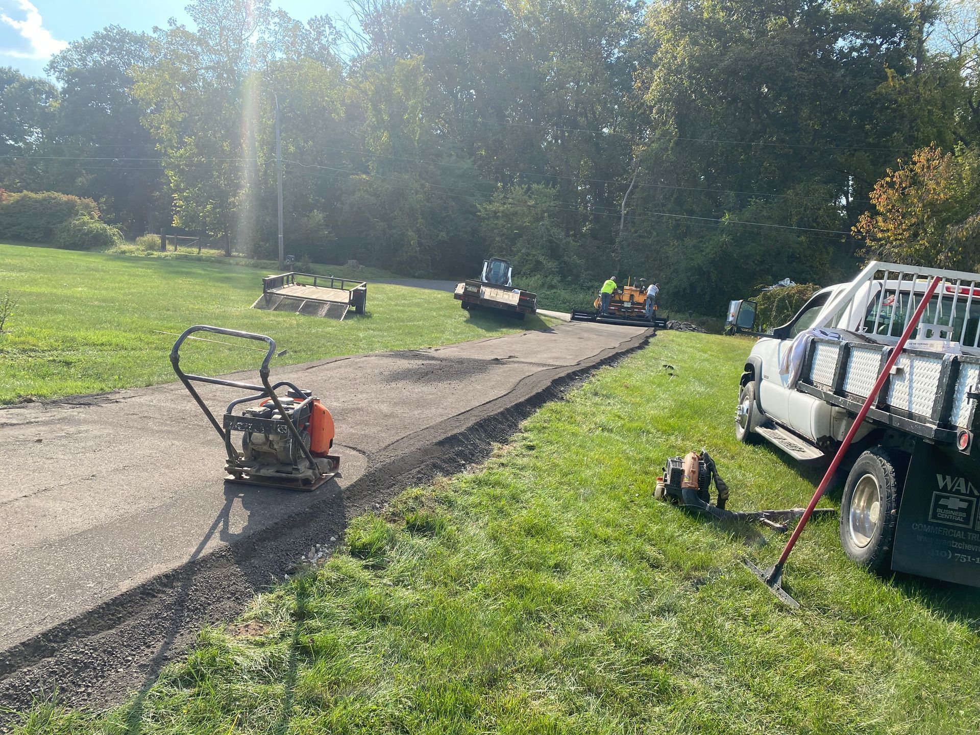 Asphalt driveway being resurfaced by a work crew; machinery and truck on green lawn.