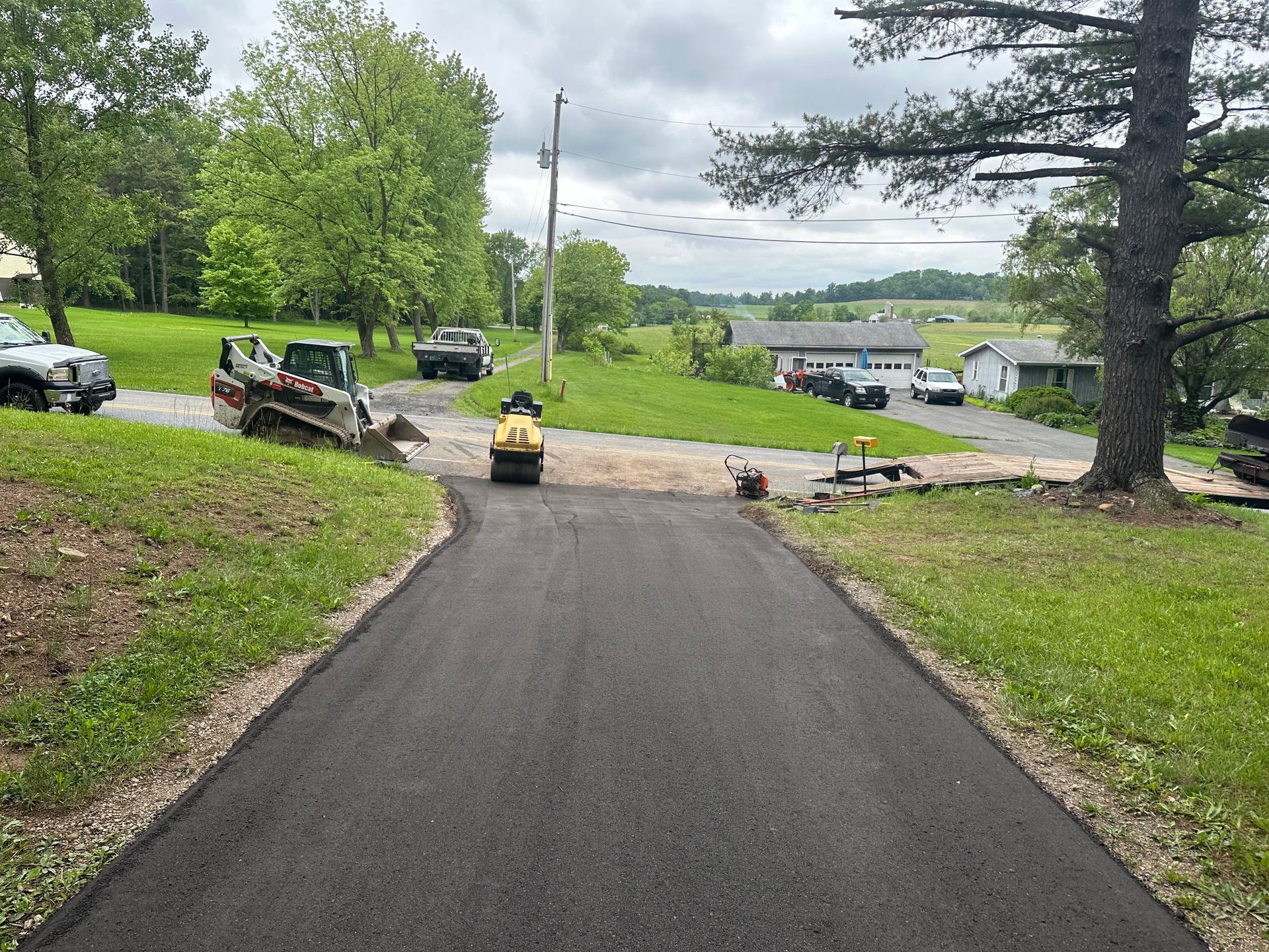Asphalt driveway being rolled by a machine; surrounding grass and trees, rural area with parked vehicles.