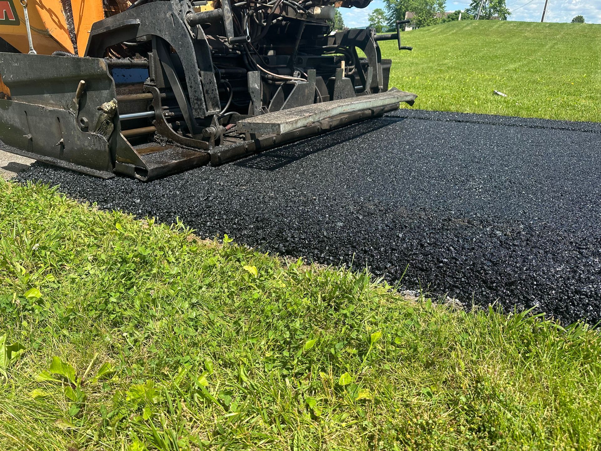 Asphalt paving machine laying fresh black asphalt on a road, next to green grass.