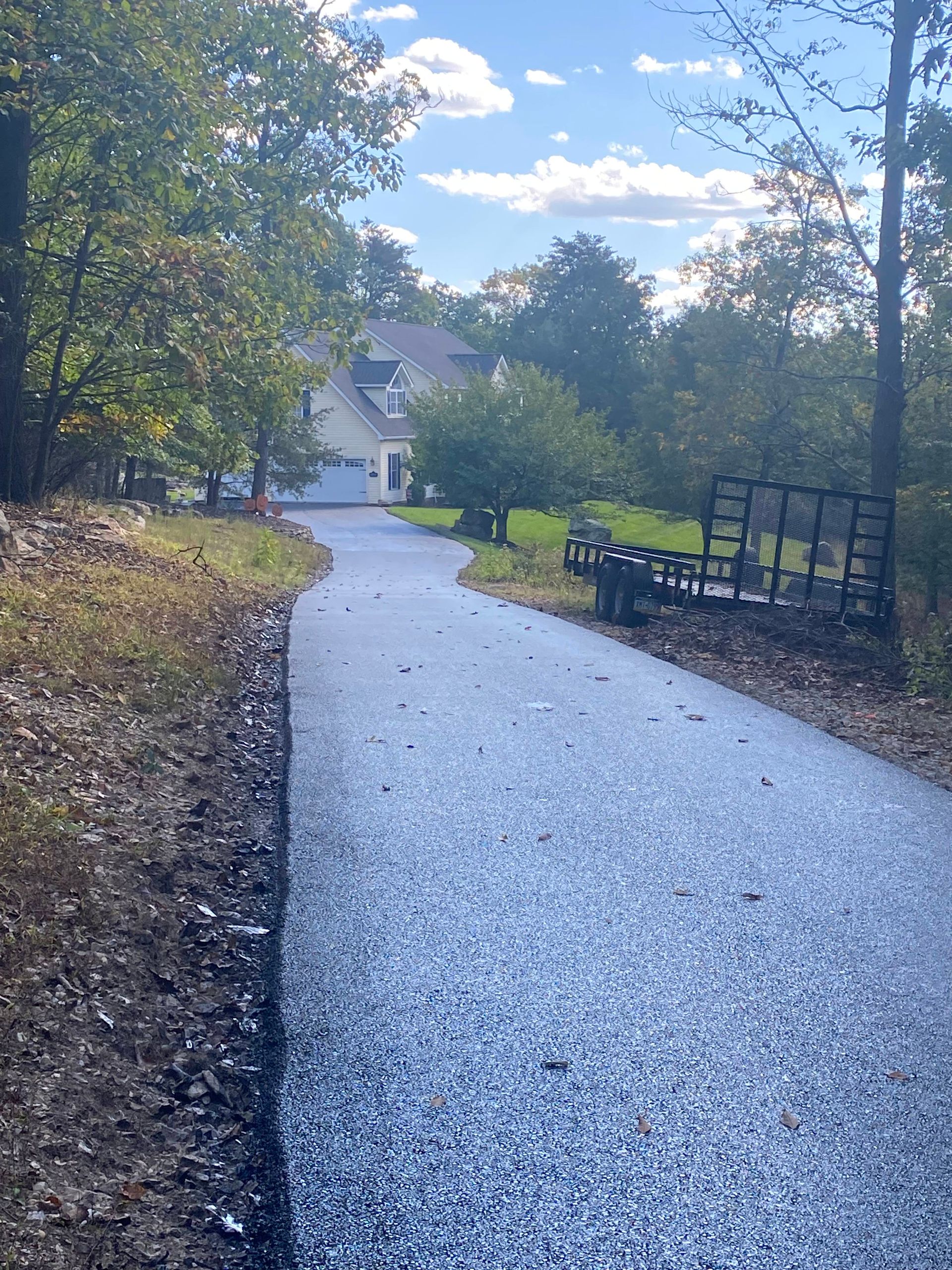 Paved driveway winding uphill toward a house, flanked by trees and foliage under a blue sky.
