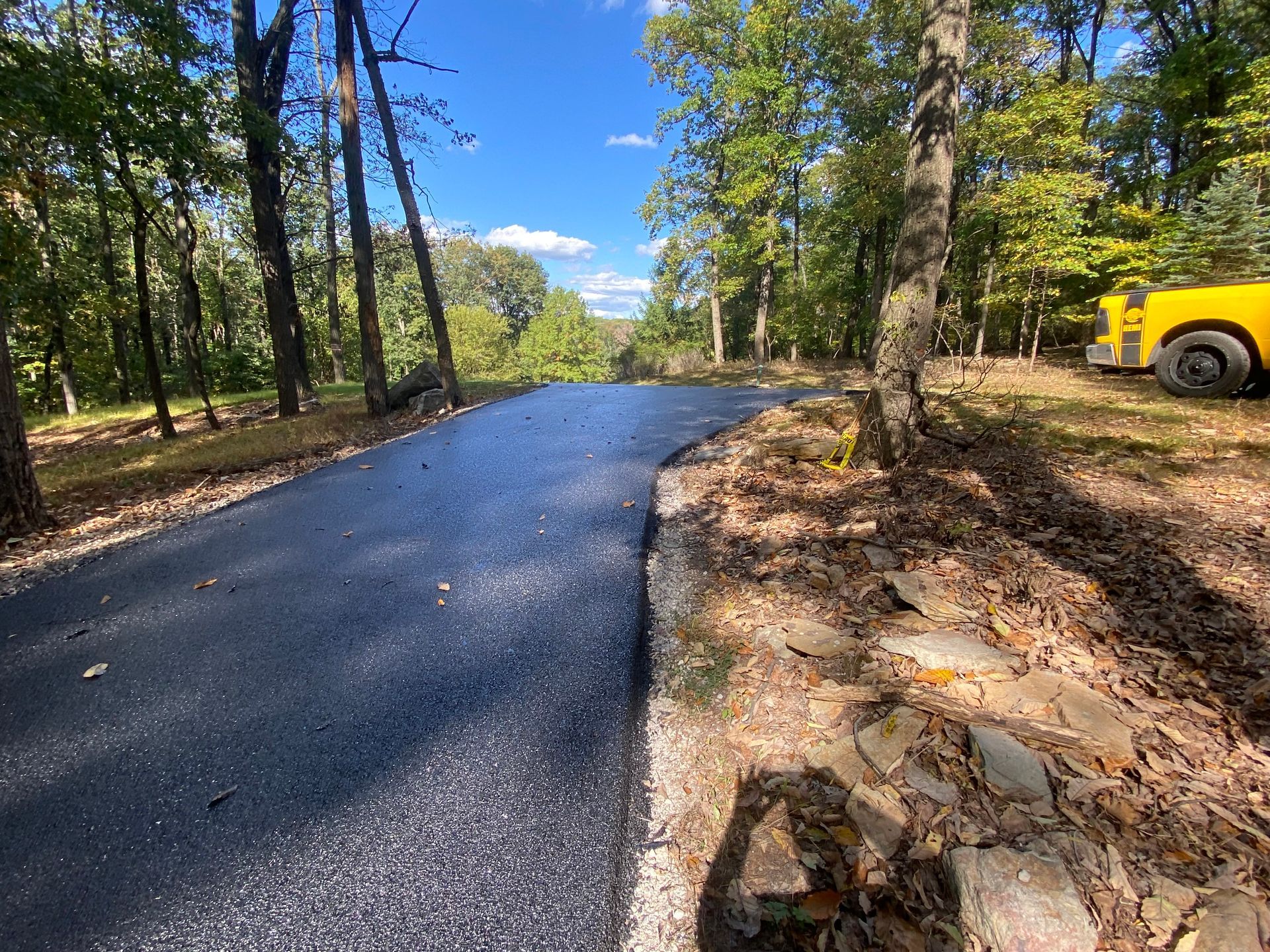 Paved road curves uphill through wooded area. Yellow vehicle parked on right. Sunny day.