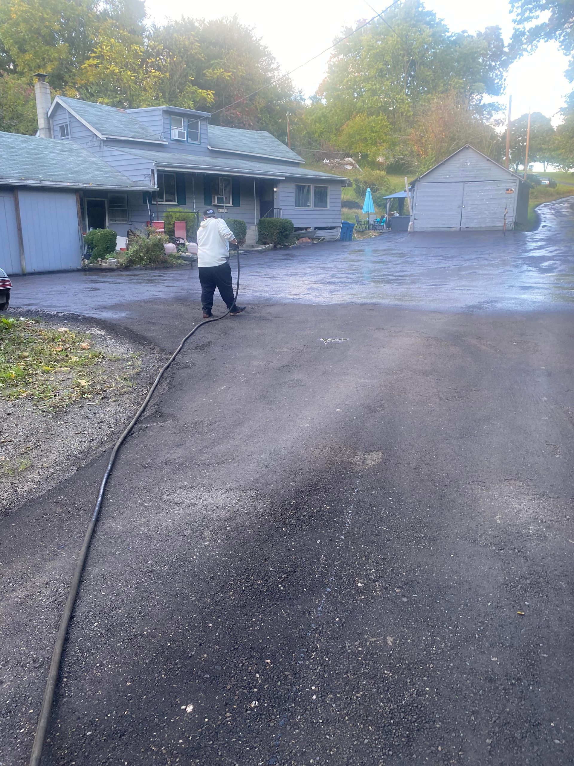 Person spraying a dark driveway near a gray house and a shed.