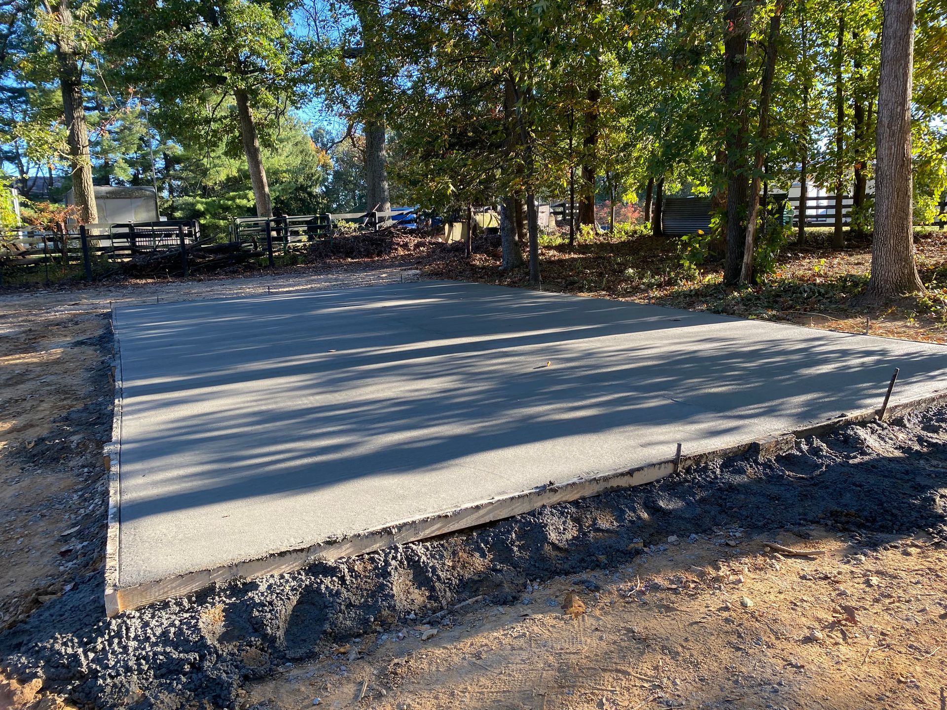 Newly poured concrete pad, gray, bordered by gravel. Trees and fence in background, outdoors.