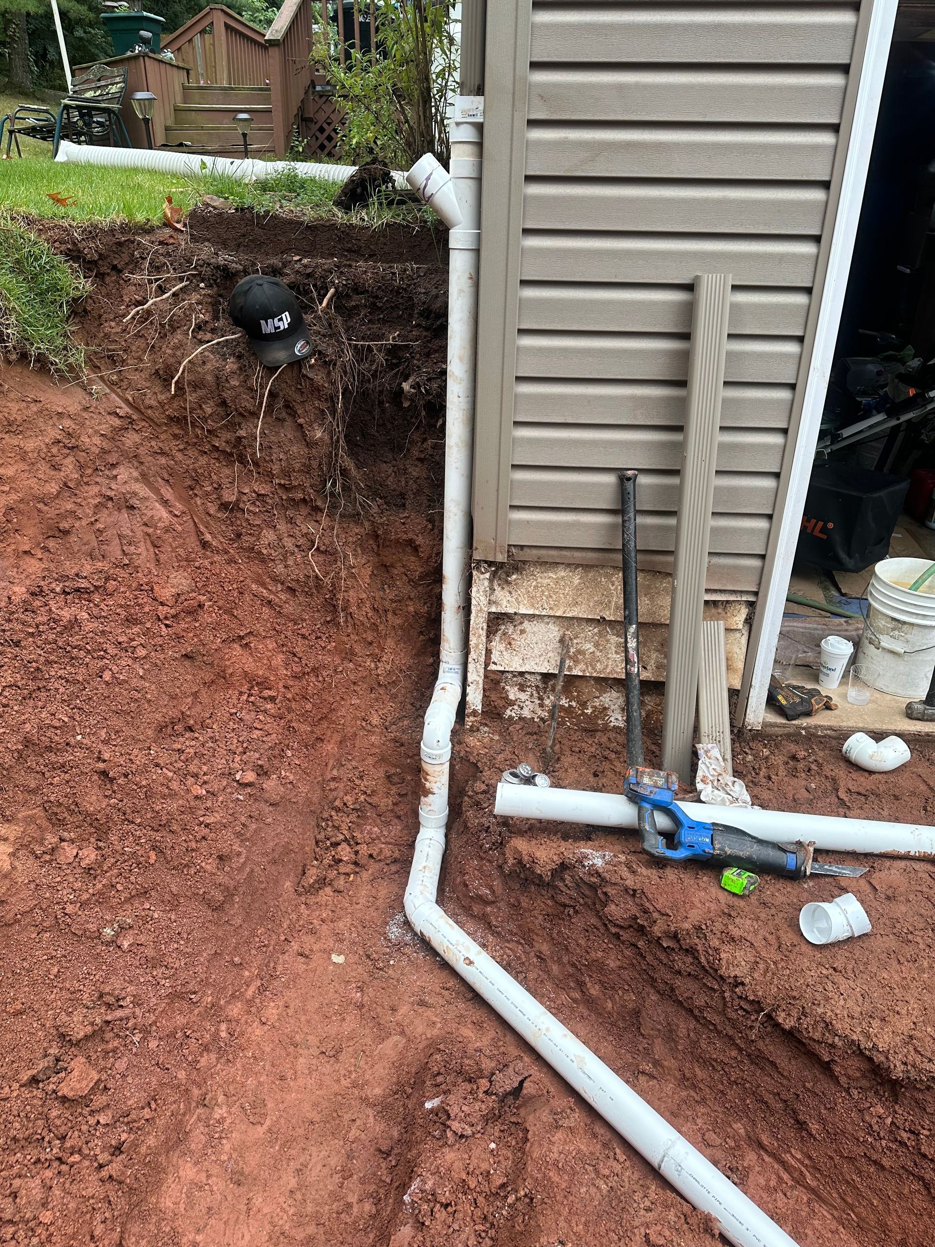 White drainage pipes being installed near a building's foundation and reddish-brown soil.