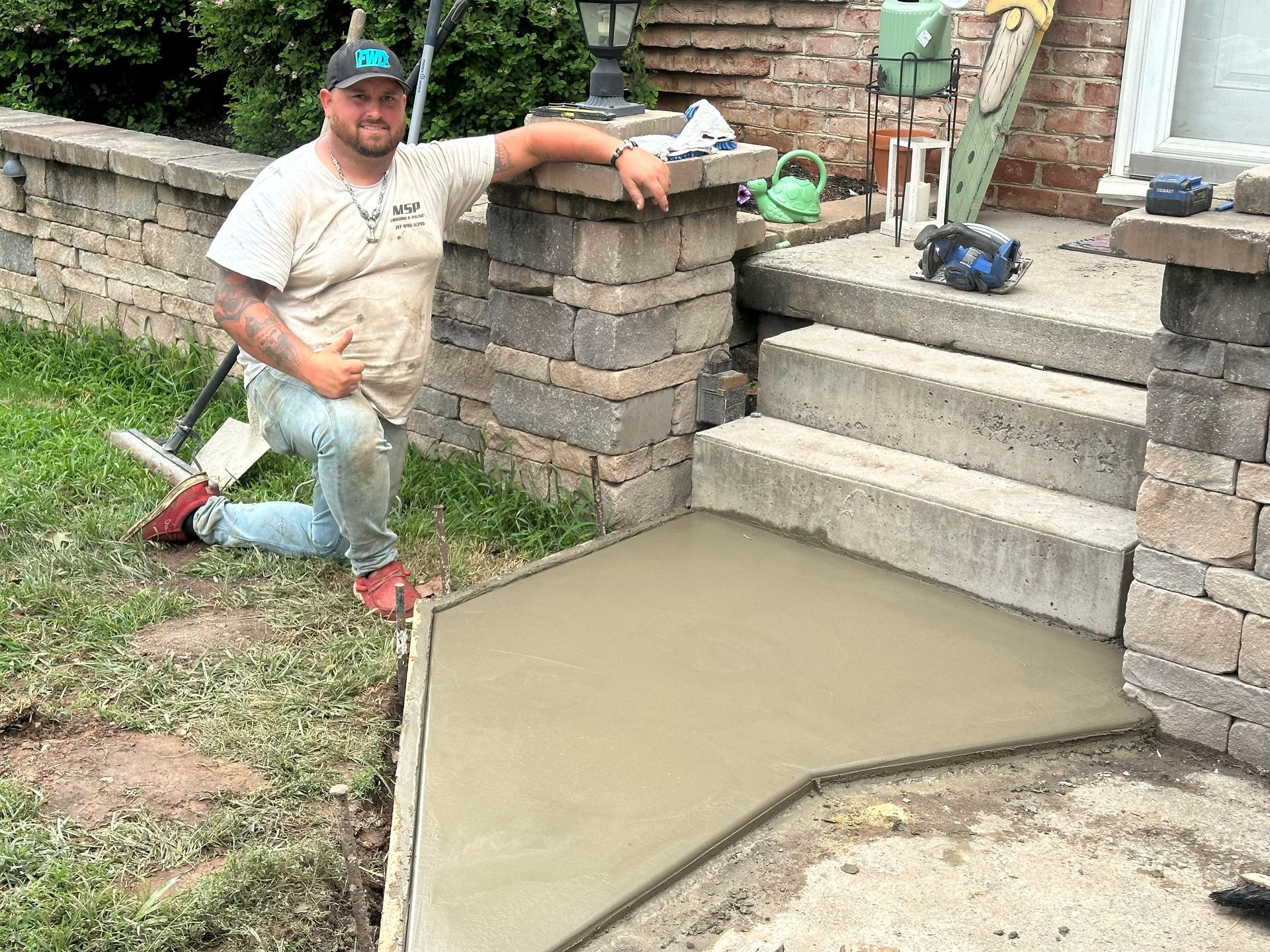 Man kneeling beside newly poured concrete pathway and steps, posing near stone pillars and wall.