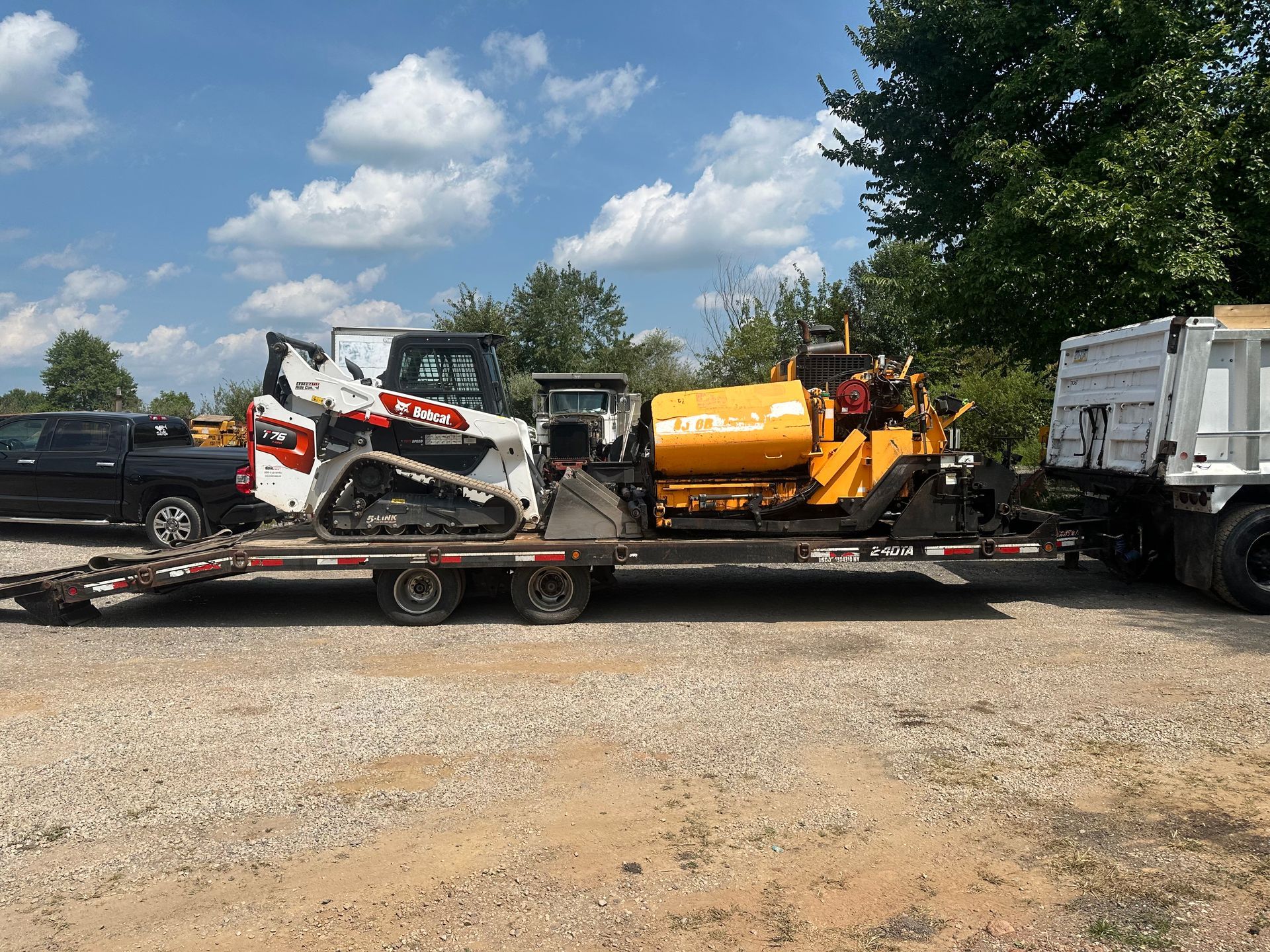 Construction equipment on a flatbed trailer: skid steer, excavator, asphalt paver, and dump truck under a blue sky.