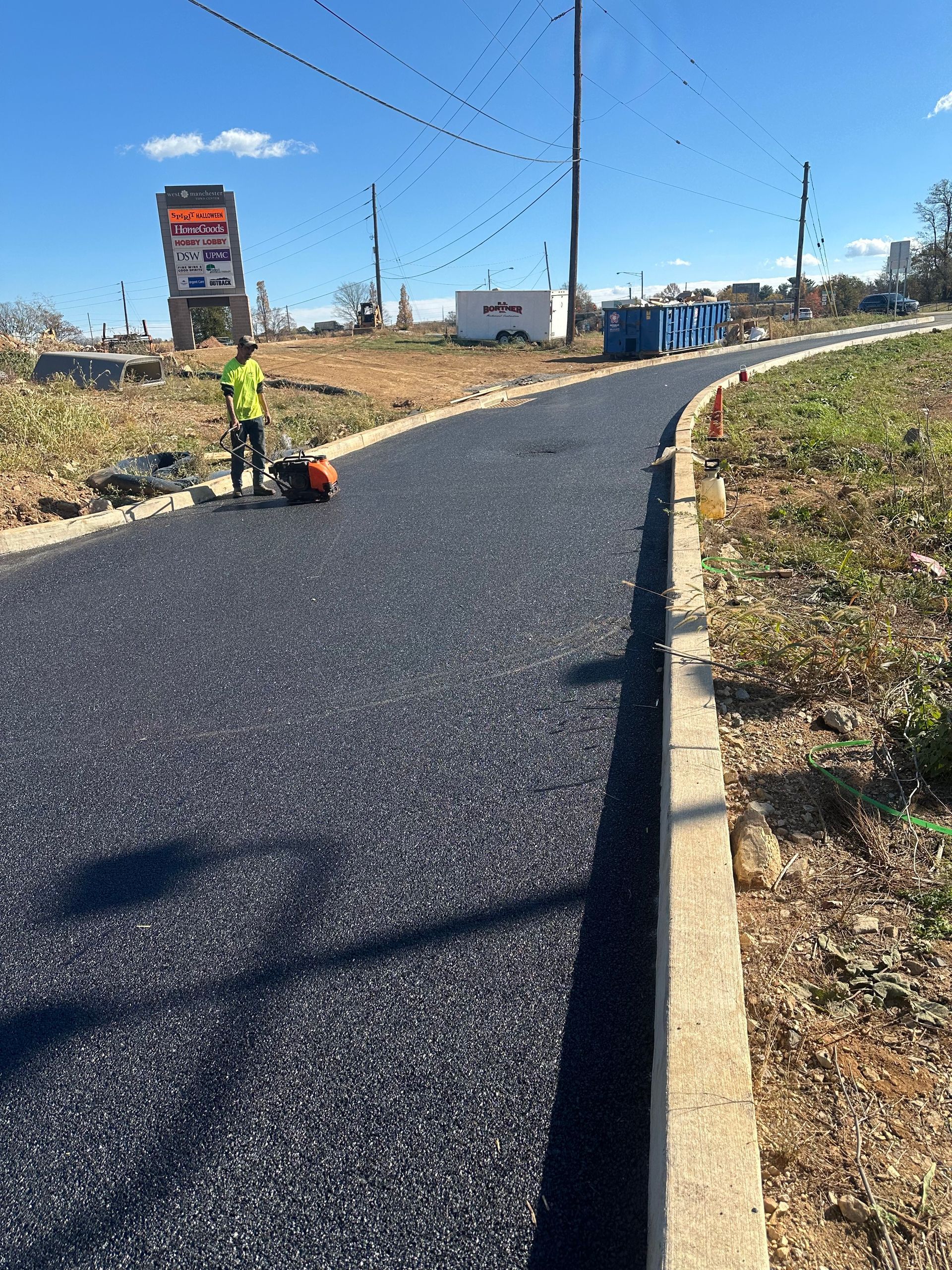 Man compacting asphalt on a newly paved road with a concrete curb in a construction zone.