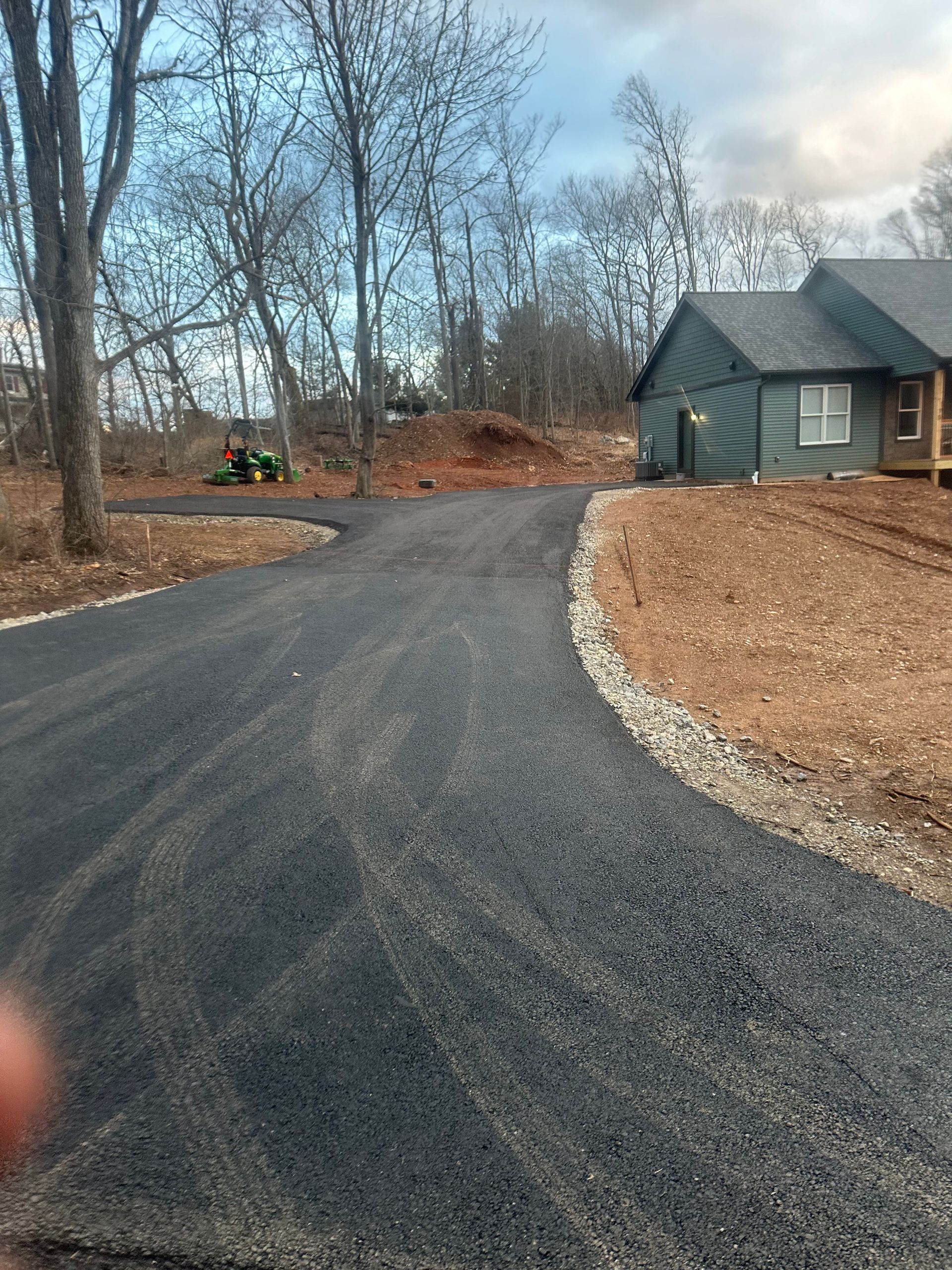 Newly paved asphalt driveway curves toward a house with green siding, in a wooded area.