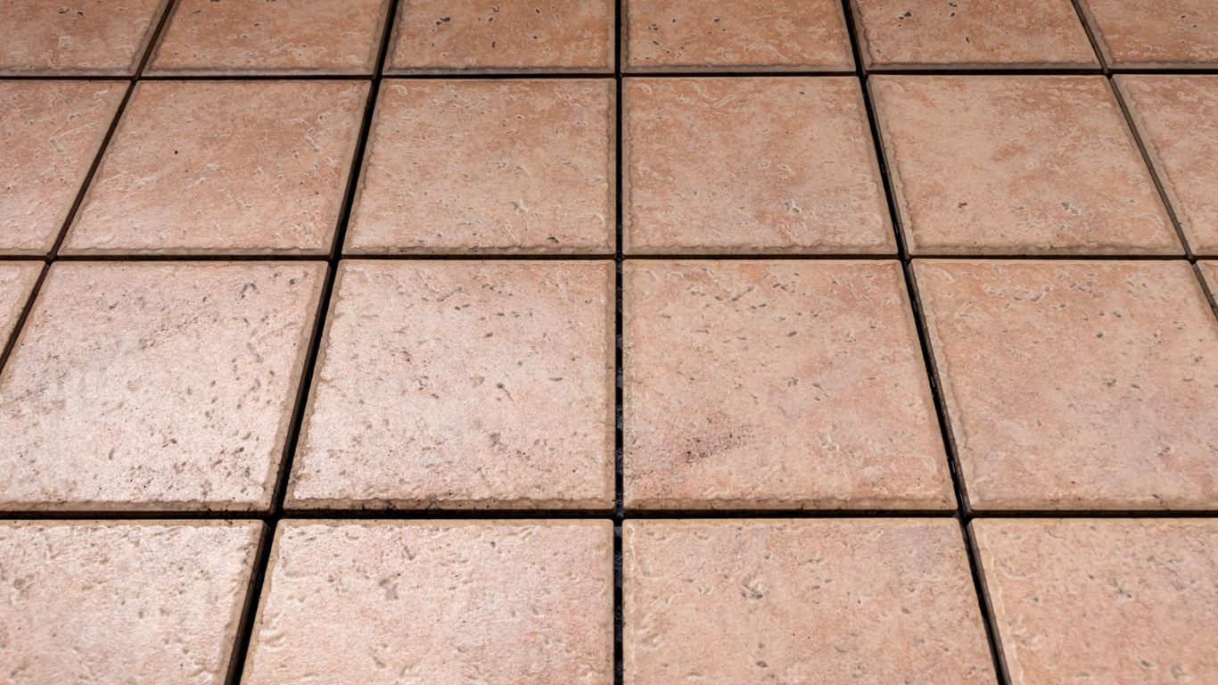 A close-up view of light brown, square ceramic floor tiles with dark grout lines, set in a repeating grid pattern.