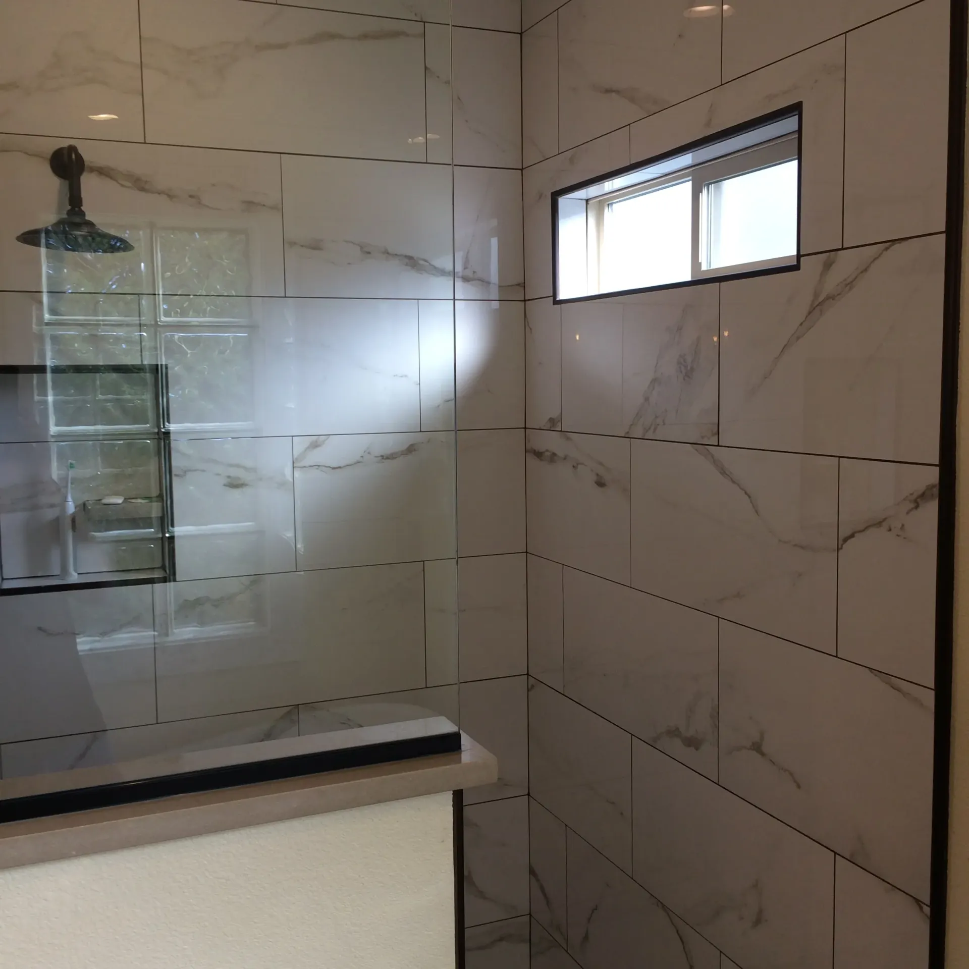 A shower stall with white marble-patterned tile, a small rectangular window, and a dark-finished shower head.