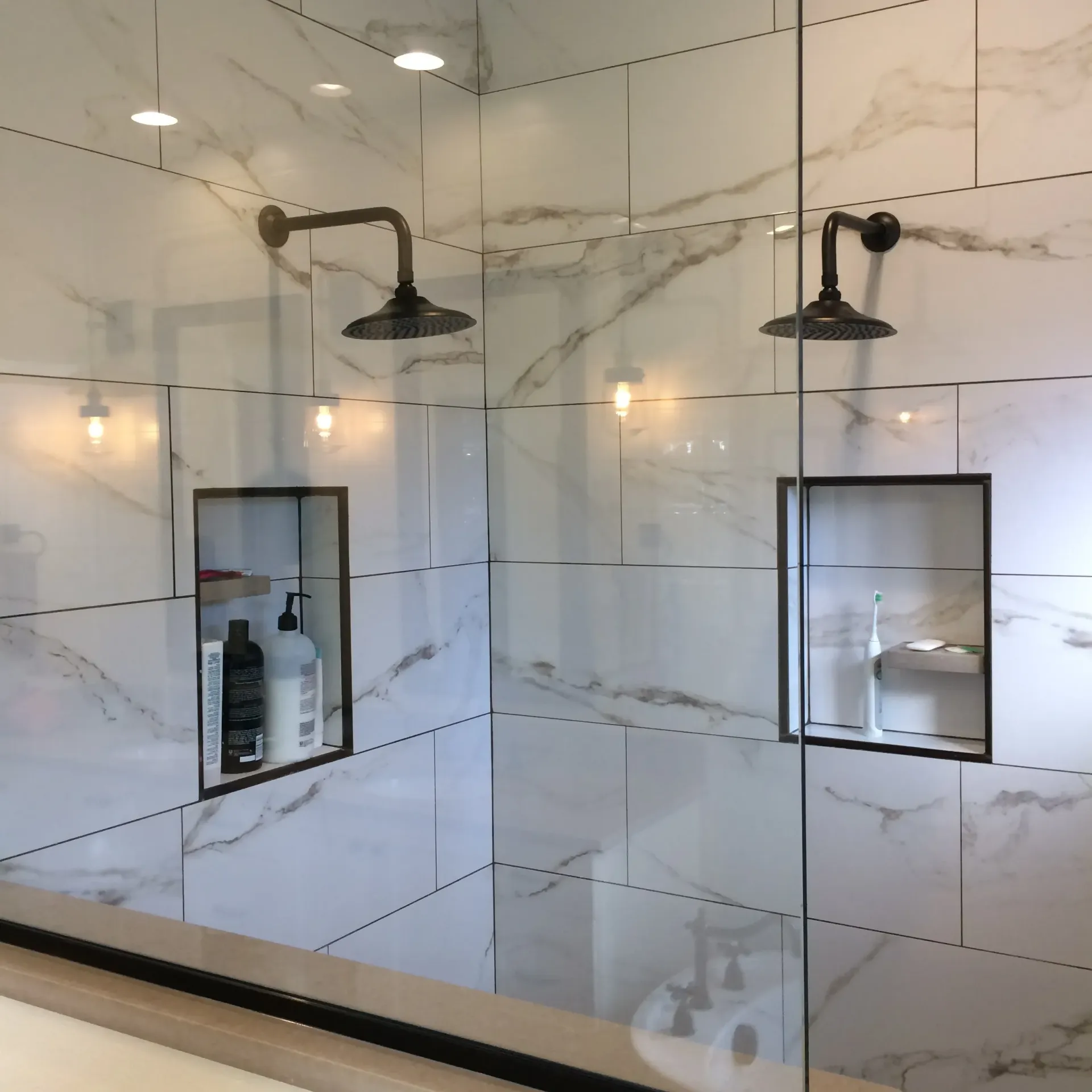 A modern walk-in shower with white marble-patterned tiles, two dark rainfall showerheads, and two recessed wall niches.