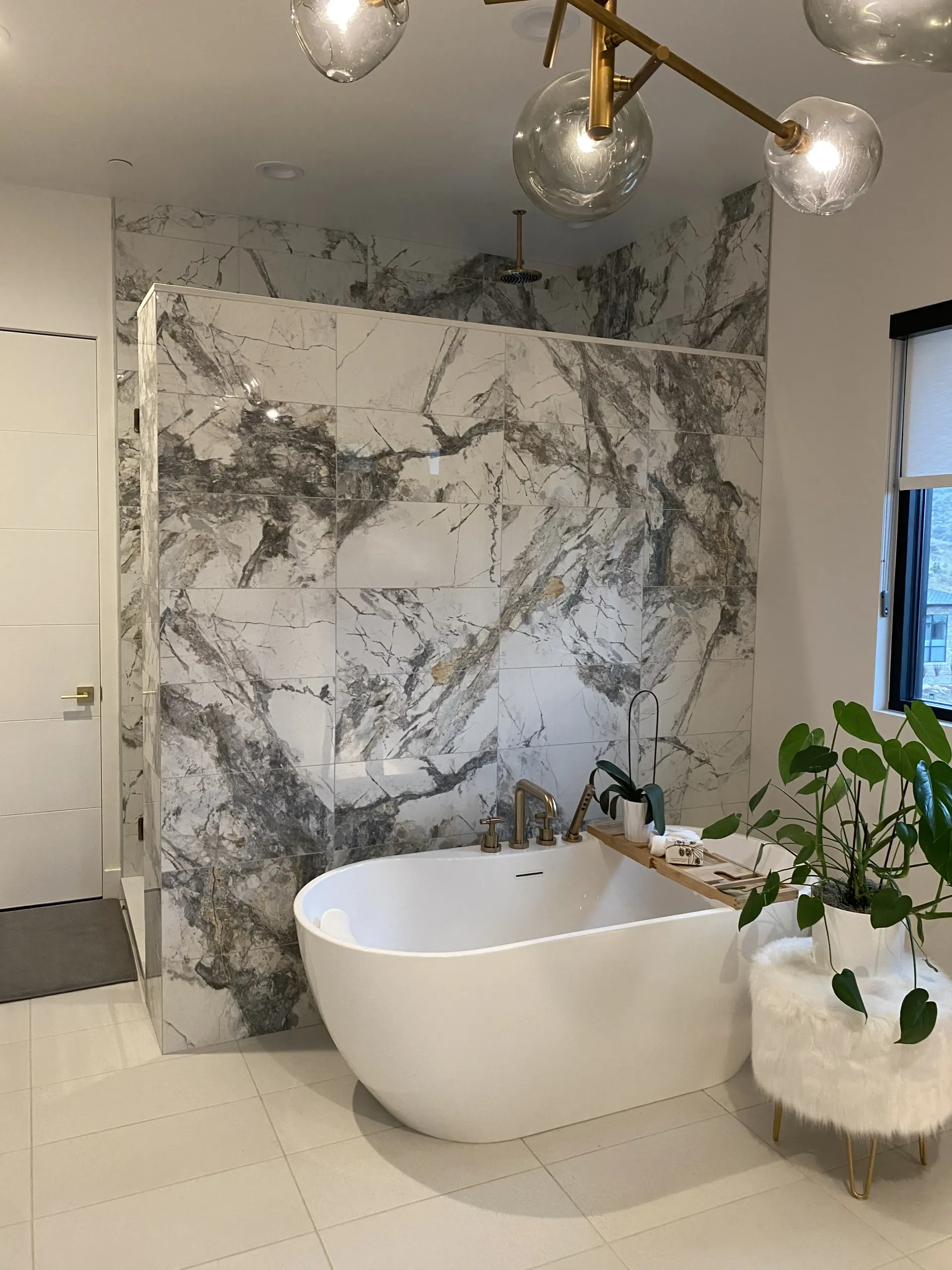 A white freestanding bathtub sits in front of a wall tiled with large, dramatic white and grey marble slabs.