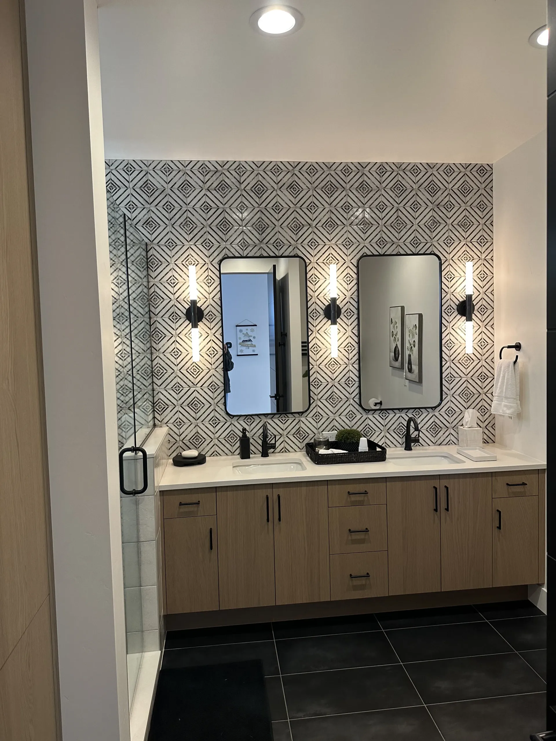 Modern bathroom with a patterned tile wall, double vanity with wooden cabinets, two mirrors, and black tiled flooring.