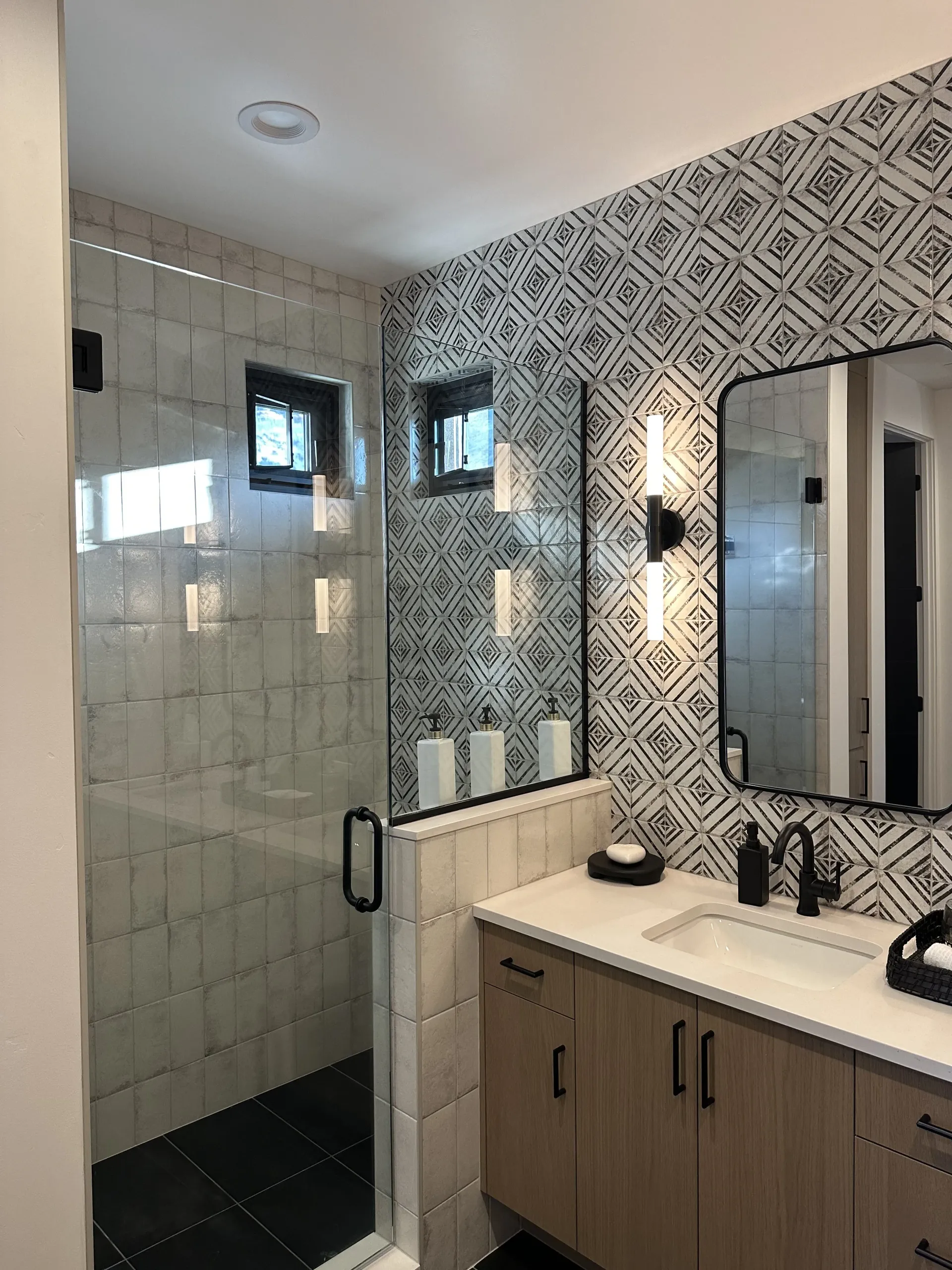 A modern bathroom with a glass-enclosed shower, wood vanity, white countertop, and black-and-white patterned wall tile.